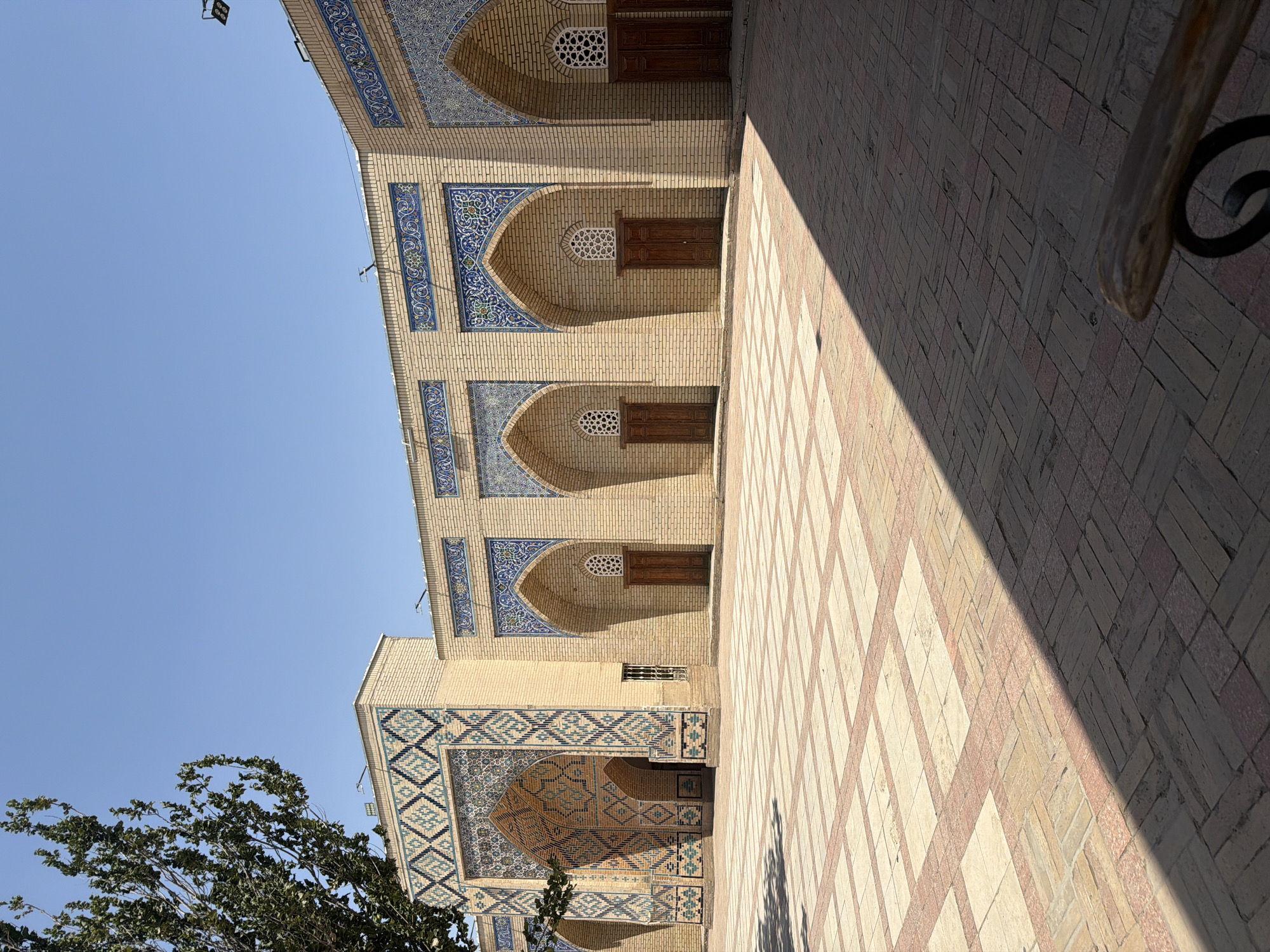 This is a wide-angle, eye-level, outdoor photograph of the exterior of a historic building complex in Zangiata, Uzbekistan. The architecture is characterized by light-colored brickwork, with intricate blue and white tile mosaics adorning the arched doorways and upper facades. Several arched doorways with wooden doors are visible along the building. The courtyard in front is paved with a pattern of rectangular stones and brickwork, partially covered by a large shadow cast by an unseen object. A portion of a leafy green tree is visible on the left side of the frame. The sky is clear and blue. There are no people visible in the scene.