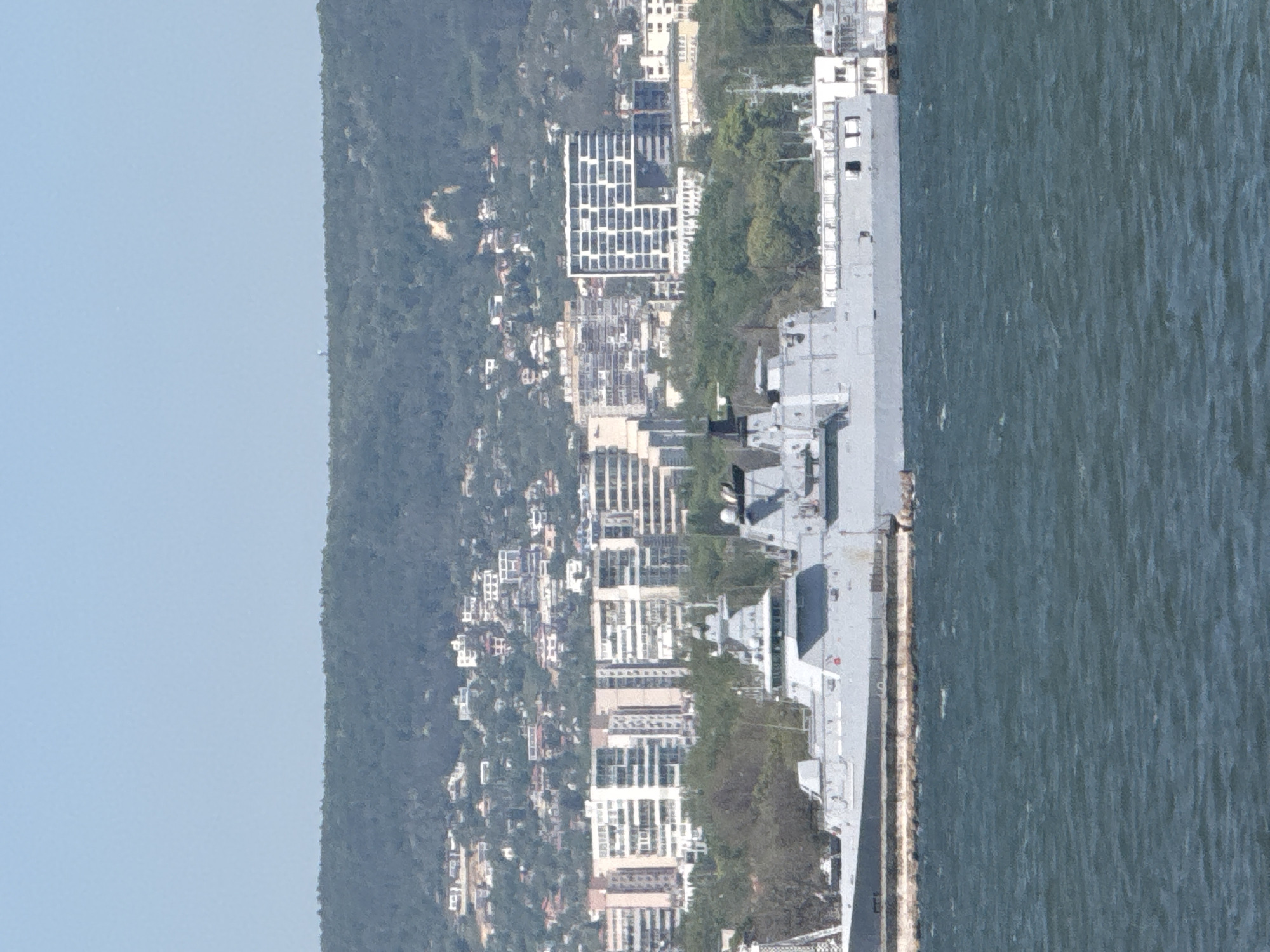 The image shows a cityscape adjacent to a body of water, likely the Black Sea, with a large grey naval vessel docked along the coast. The city is densely populated with residential and commercial buildings, and a forest-covered hillside forms the backdrop. The vessel appears to be a type of landing platform dock or assault ship, evident from its flat top and the presence of possible helicopter landing spots. The environment suggests a clear, daytime setting. Given the context of Varna, Bulgaria, the location is likely somewhere along the coastline where naval vessels dock.