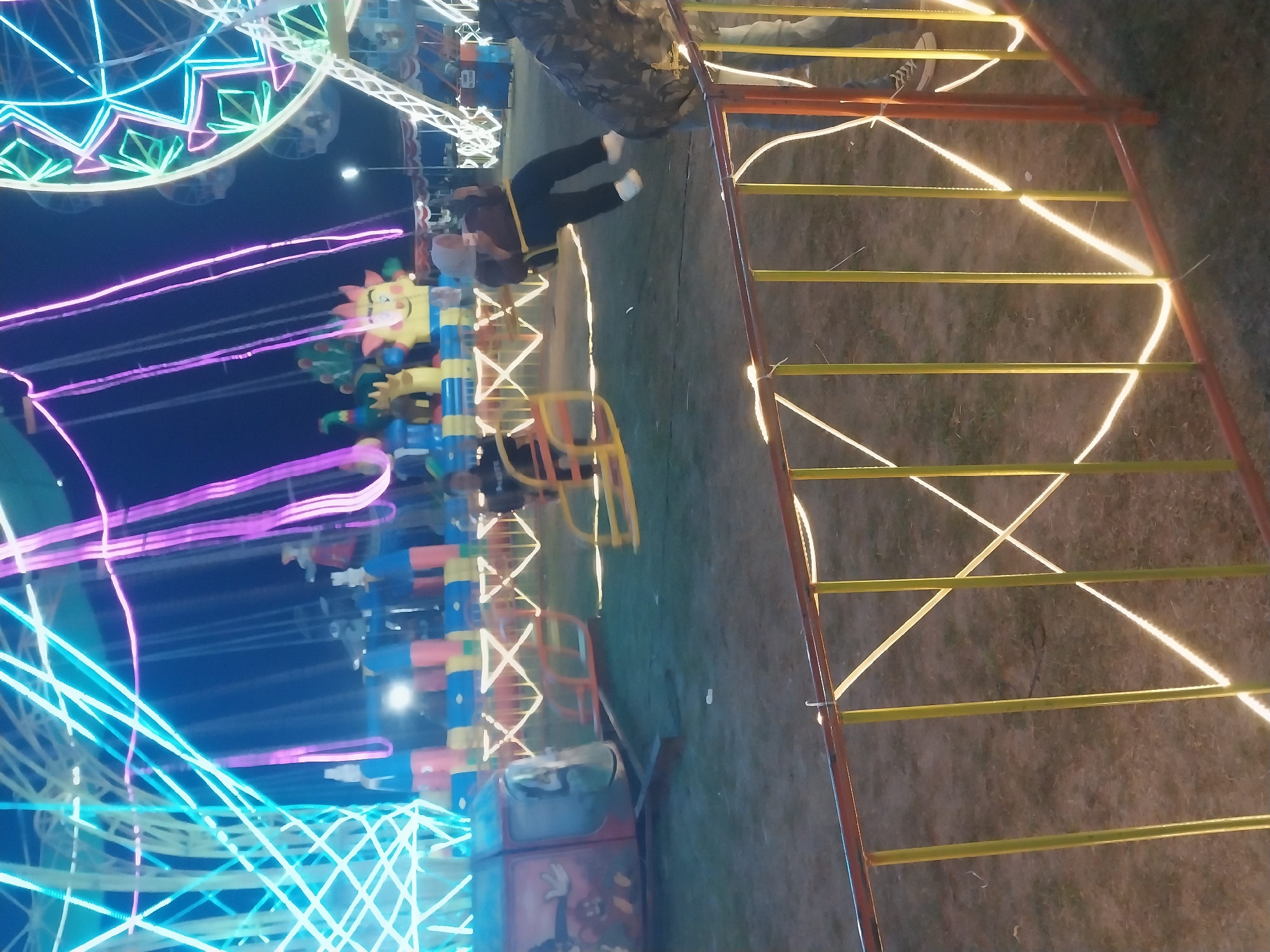 The image depicts a night scene at a carnival or amusement park in Jiwut, Indonesia. The foreground features a railing with yellow vertical bars and orange horizontal bars, illuminated by a string of warm white lights. Beyond the railing, the ground is covered in dry grass and dirt. In the middle ground, a swing ride is in operation, with several people on it. One person, wearing a hijab and a brown top with black pants, is swinging. Other amusement park rides are visible in the background, including a Ferris wheel and a carousel-like structure adorned with bright blue, pink, and white lights. The overall atmosphere is festive, illuminated by the colorful lights of the rides against the dark night sky.