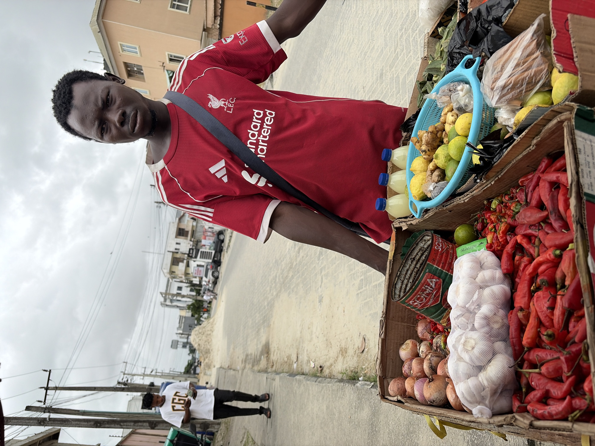 In Igboefon, Nigeria, a young Black man wearing a red Liverpool FC jersey and a dark crossbody bag stands beside a cart filled with fresh produce. The cart contains boxes overflowing with red chili peppers, onions, and mesh bags of garlic. A blue basket holds lemons, limes, and ginger. Behind the man, a street scene unfolds with several buildings, power lines, and a pile of sand. Another individual, dressed in a white t-shirt with the letters "CGC" and dark pants, is seen walking on the sidewalk in the background, holding a wad of cash. The sky is overcast, suggesting a cloudy day.