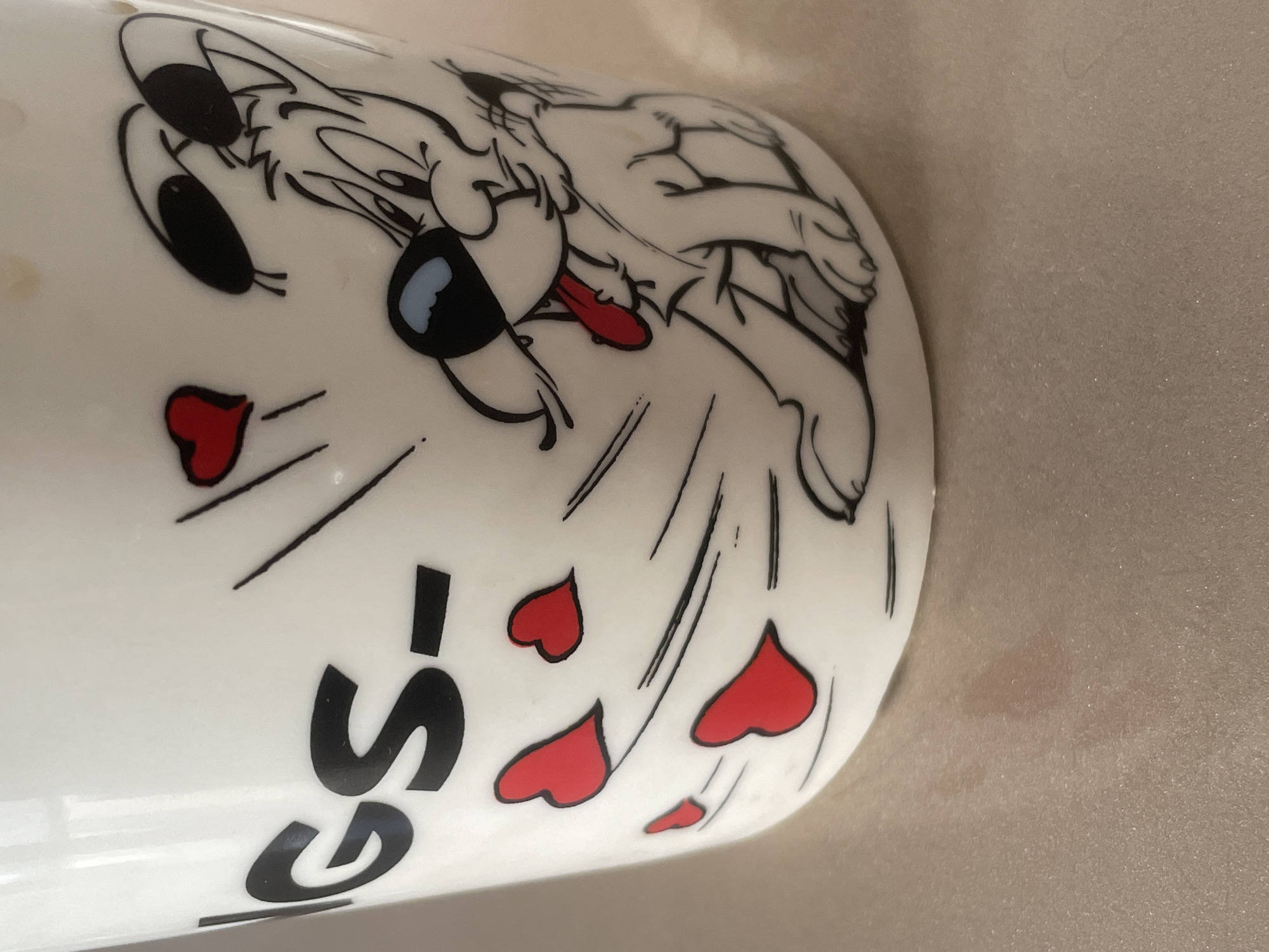The image is a close-up shot of a white ceramic mug with a cartoon dog illustration. The mug is decorated with black lines and red hearts, suggesting a whimsical or affectionate theme. On the left side of the mug, black lettering is partially visible, reading "IGS-". The cartoon dog is depicted with a large head, big ears, and a red tongue sticking out, surrounded by red hearts. The mug is placed on a textured surface, possibly a table, which appears to have a slight sheen. There are some subtle pinkish stains on this surface in front of the mug. The lighting suggests an indoor environment with ambient light, casting gentle reflections on the mug and the surface. The background is out of focus, showing a soft, blurred light color. The image does not contain any information or visual cues to suggest a location in Tashkent, Uzbekistan.