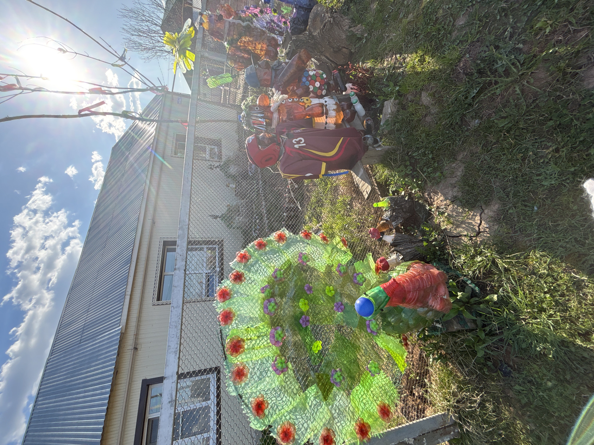 The image shows a yard or garden scene in what appears to be the city of Фоки, Russia. A weathered, two-story building with a corrugated metal addition stands behind a chain-link fence. Sunlight illuminates the yard, where several objects are visible. A decorative item resembling a peacock, constructed from recycled plastic bottles, sits on the ground near the fence. A maroon and yellow sports bag or duffel bag, marked with the number "13," leans against the fence alongside a red helmet. A pile of assorted plastic containers and debris is gathered along the fence line, possibly used as decorations or part of a larger art installation. The yard is covered in grass and weeds.