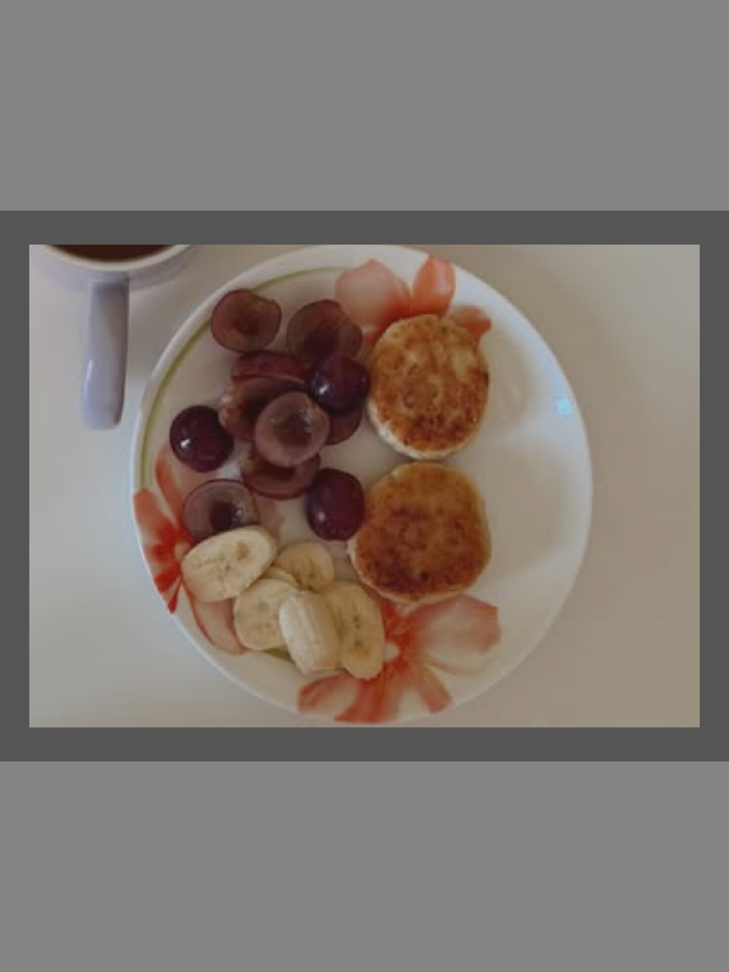 A close-up, overhead shot shows a plate of breakfast food. Two golden-brown pancakes are placed side-by-side on the right of the plate. To the left of the pancakes are sliced grapes and banana. The plate itself is white with a floral pattern of red and orange flowers around the rim. A white mug with a dark liquid inside is partially visible in the upper left corner of the frame. The background is a plain white surface.
