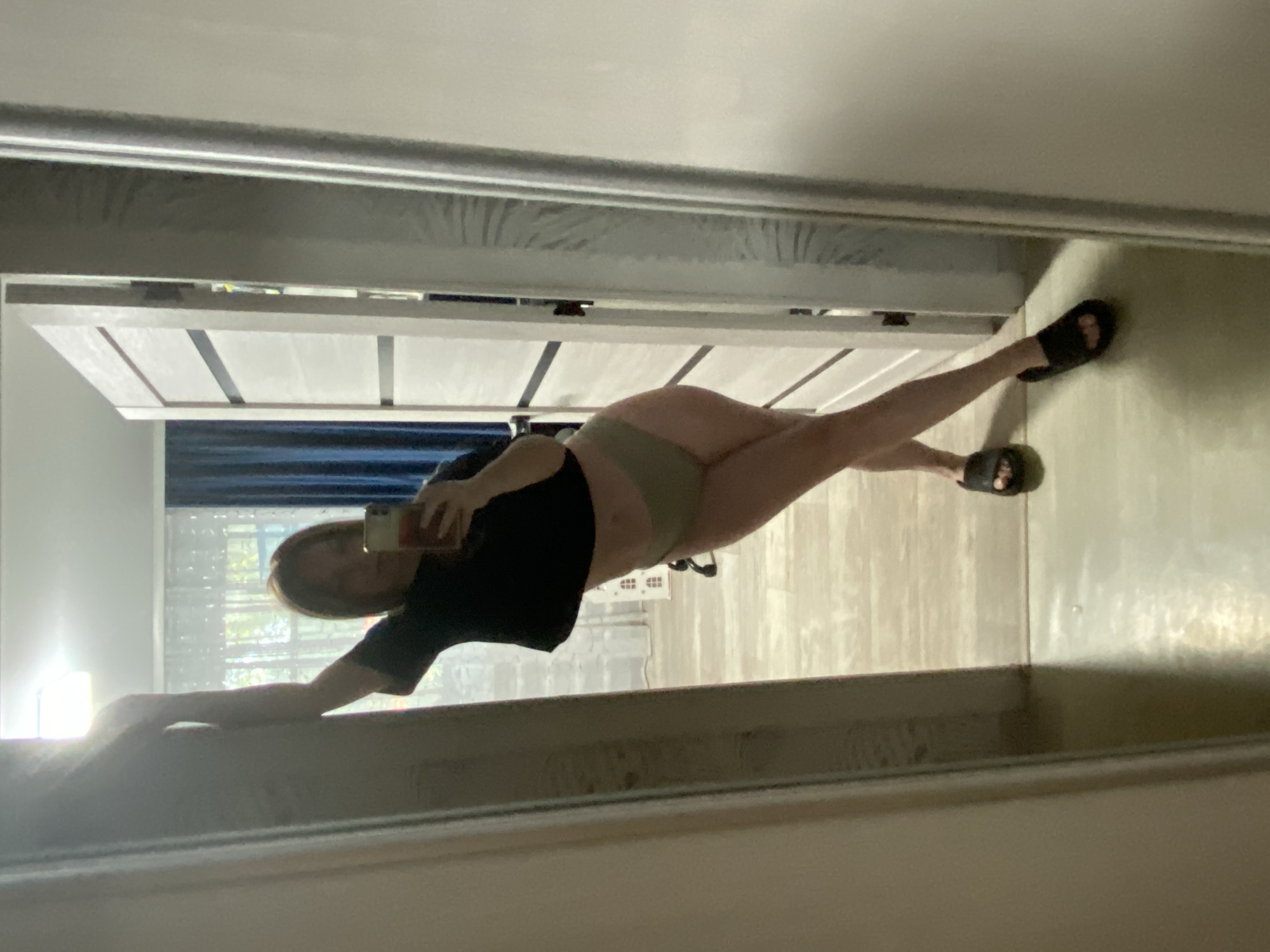 A woman is taking a selfie in a mirror. She is wearing a black shirt and tan bottoms. She is wearing sandals on her feet. The background includes a door, curtains, and a wall. The location cannot be determined to be Noviy, Russia, or any specific place, based on the image alone.