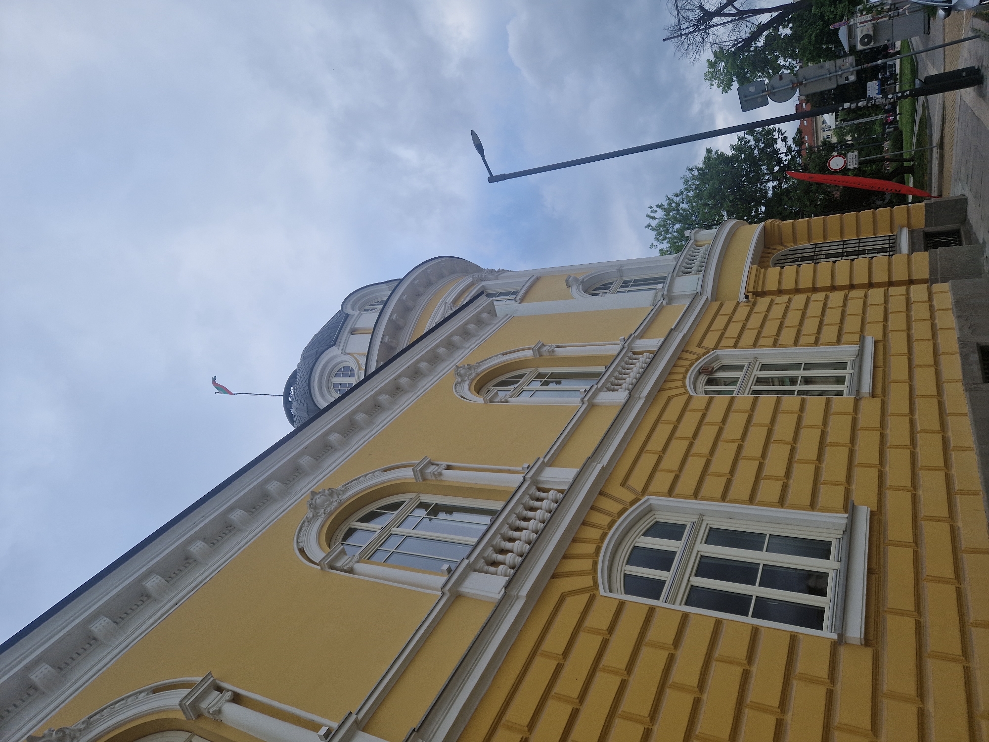 The image shows a yellow building in an unknown location, featuring a patterned brick facade, arched windows with white frames, and ornate white detailing along the building's upper sections. A domed roof is visible at the top, with a flag on a pole extending from it. A modern street light is situated in the upper-left, and a street with trees, signs, and pedestrian crossing is partially visible on the right side of the image. The sky is overcast.