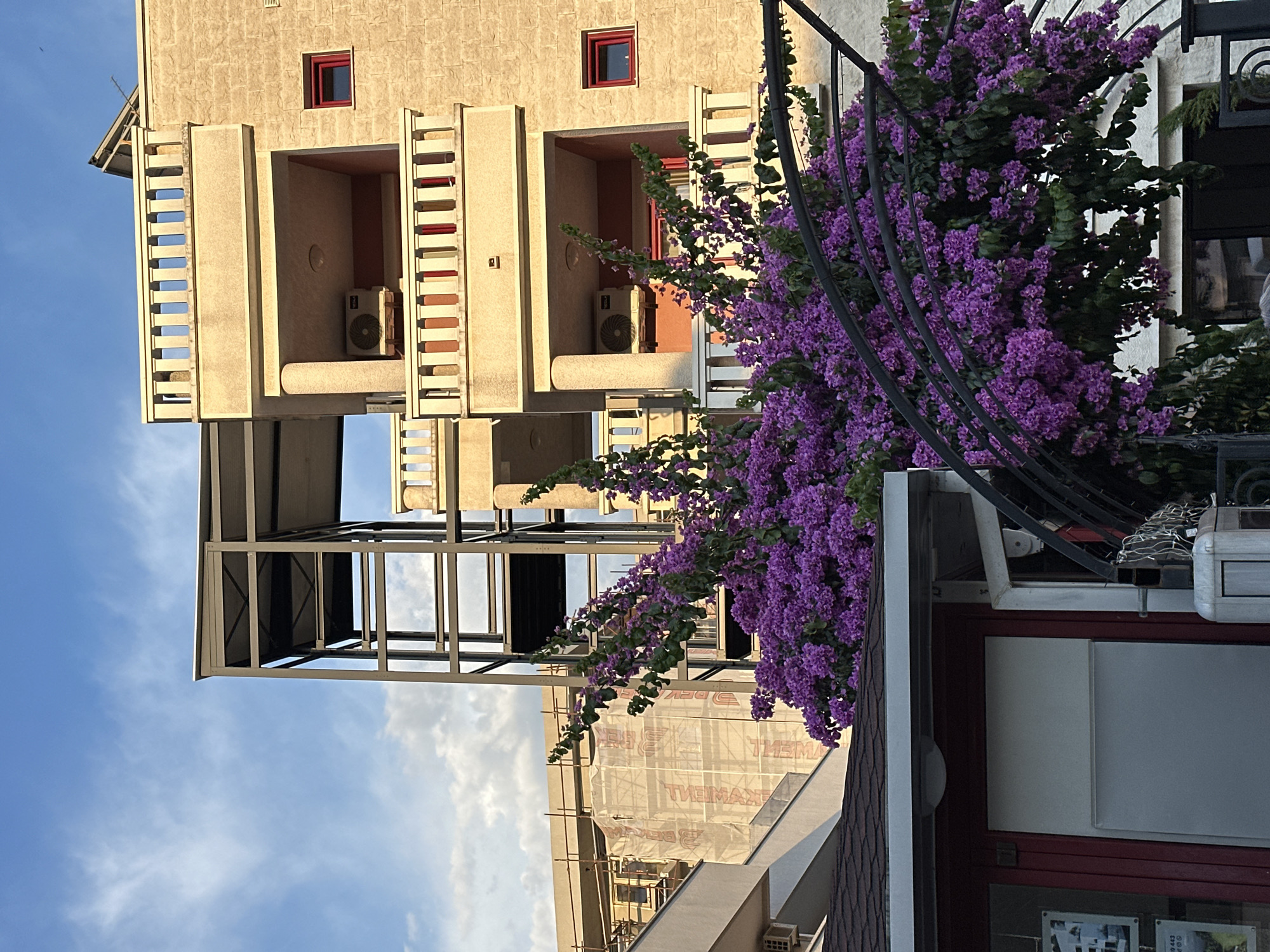 The image captures a street view in Budva, Montenegro, during the late afternoon. In the foreground, a low-rise building with a dark-tiled roof and red-framed doors is partially visible, with an outdoor seating area covered by a black metal archway heavily adorned with vibrant purple bougainvillea flowers. Behind this, a multi-story building constructed with light-colored textured stone features balconies with white railings. One balcony shows an air conditioning unit and a small red-framed window. Further in the background, a more modern structure with a dark metal frame rises, partially obscuring a building covered in white construction sheeting and scaffolding. The sky above is a clear blue with scattered white clouds, and the lighting suggests the golden hour. There are no people visible in the image.