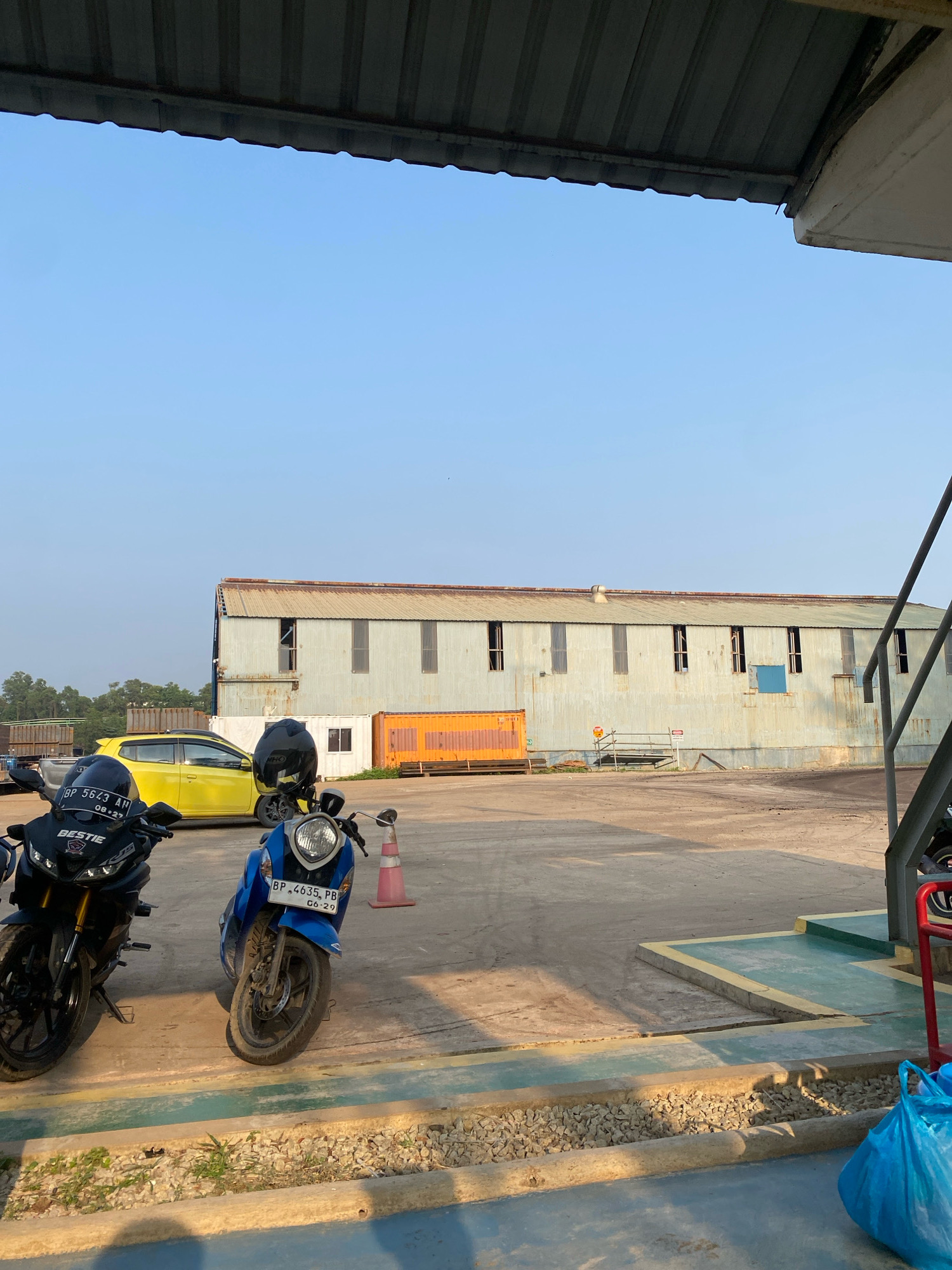 The image depicts an outdoor scene, likely in Batam, Indonesia, with a clear blue sky overhead. In the foreground, two motorcycles are parked, one black and one blue, with their license plates visible. Behind the motorcycles, a bright yellow hatchback car is parked. A large, corrugated metal building with multiple windows stands in the background. Orange and white shipping containers are also visible near the building. The ground is paved with concrete and gravel. A blue plastic bag is on the right side of the image. The overall impression is of an industrial or storage area.
