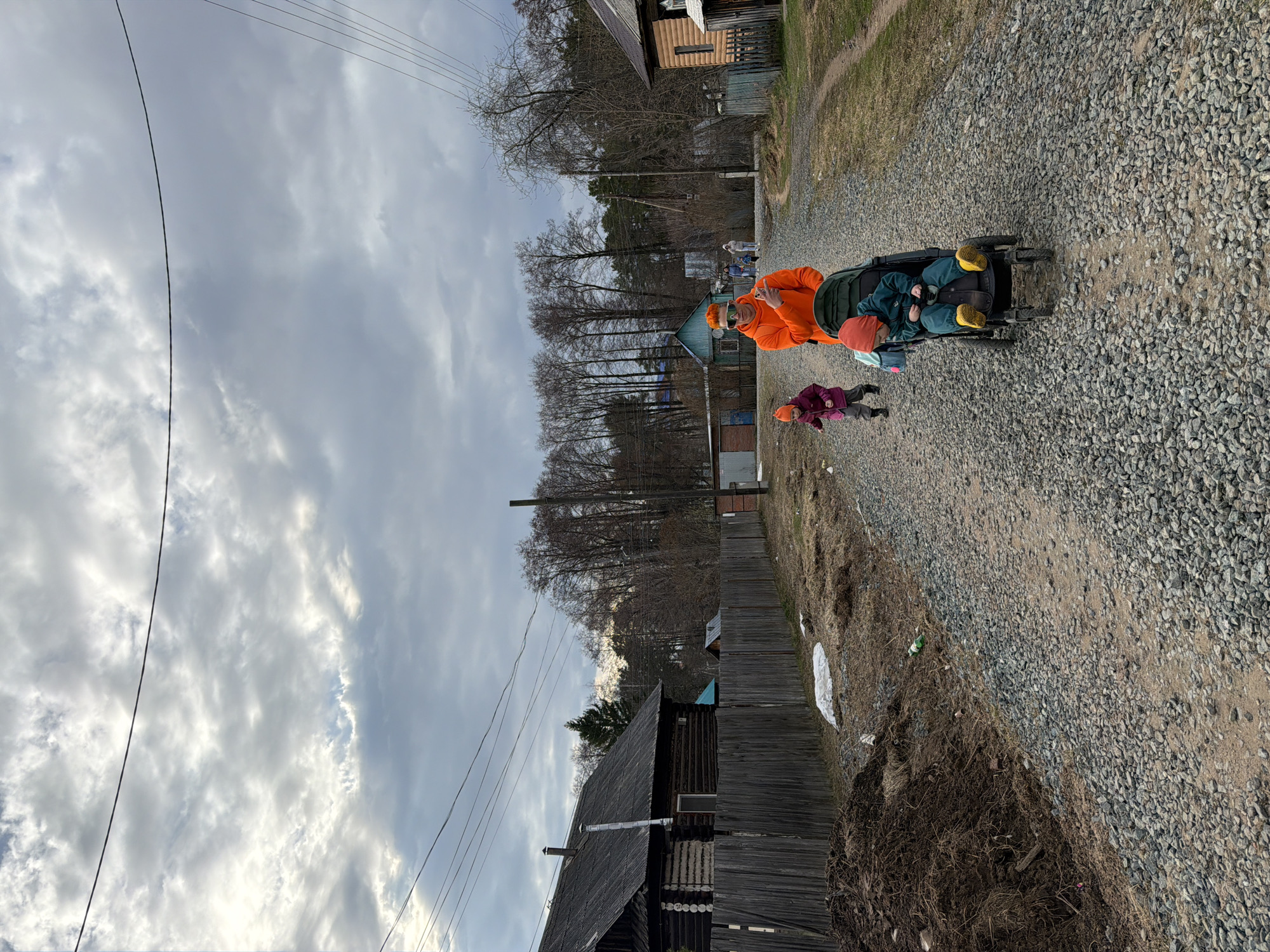 The image captures a scene along a gravel path in a rural or semi-rural area of Chaikovsky, Russia, likely during late winter or early spring.
In the mid-ground, an adult, wearing a bright orange jacket and sunglasses, pushes a dark-colored stroller. A child is seated in the stroller, dressed in a dark green or blue outfit, a red hat, and distinctive bright yellow boots. To the left of the stroller, another child, wearing a purple jacket and a red hat, walks alongside. The group appears to be on a casual outing.
The path itself is composed of grey gravel, with a visible dirt track running through it. To the left of the path, the ground is covered with dry, brown grass and dirt, with a few patches of white, possibly remnants of snow or debris. A sturdy wooden fence runs parallel to the path on the left. In the background, several traditional wooden houses with dark roofs are visible, interspersed with a dense line of tall, bare deciduous trees, indicating the season. Overhead, utility poles and multiple power lines stretch across the sky.
The sky is heavily overcast with a mix of grey and brighter white clouds, suggesting a cloudy day without direct sunlight. The overall lighting is diffuse. No specific text is visible in the image. The architectural style of the wooden structures and the natural landscape are consistent with a rural Russian setting.