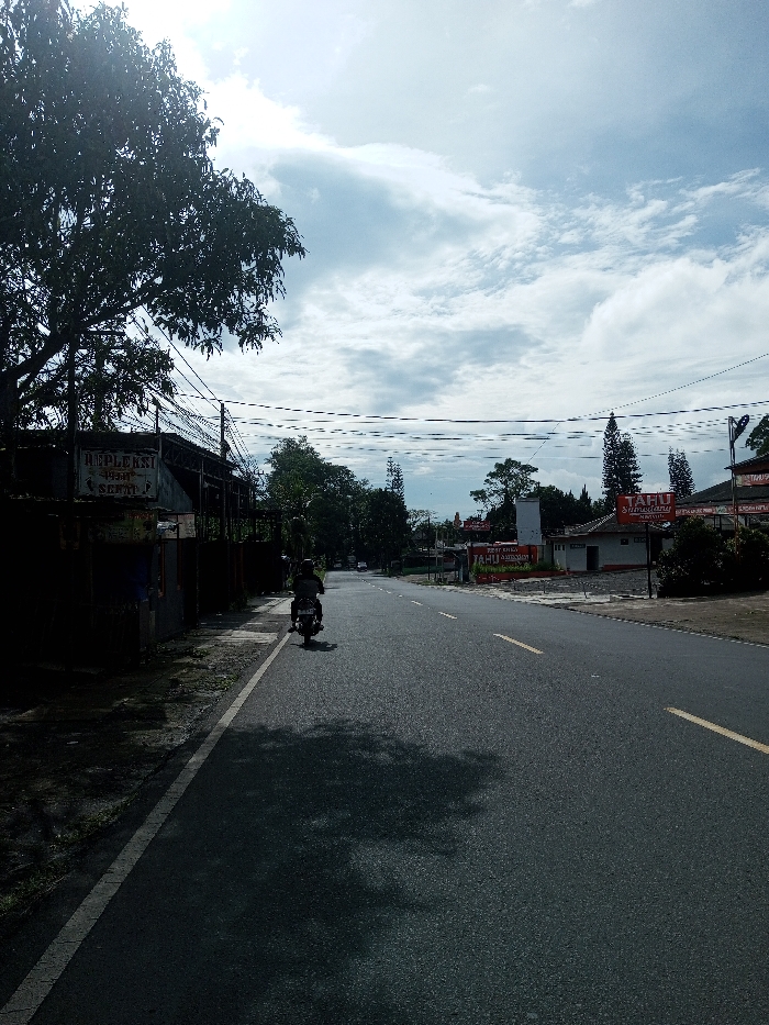 The scene depicts a daytime view of a paved two-lane road in Domphoog_Dungkarchhoeling, Bhutan, under a sky with scattered white clouds. The road, marked with a dashed yellow center line and a solid white line on its left edge, recedes into the distance. On the left side, a large, dark-foliaged tree casts a prominent shadow across the road. Beneath the tree, a building is partially visible, featuring a sign with the word "REPLEKSI" legible on it. Numerous utility wires are strung horizontally across the upper-left and central portions of the frame. A single person, wearing a dark helmet and clothing, operates a motorcycle, traveling away from the viewer in the left lane. On the right side of the road, several buildings are visible, one prominently displaying a red sign with white text that reads "TAHU SUMEDANG". Coniferous and deciduous trees are interspersed among the buildings and along the roadside.