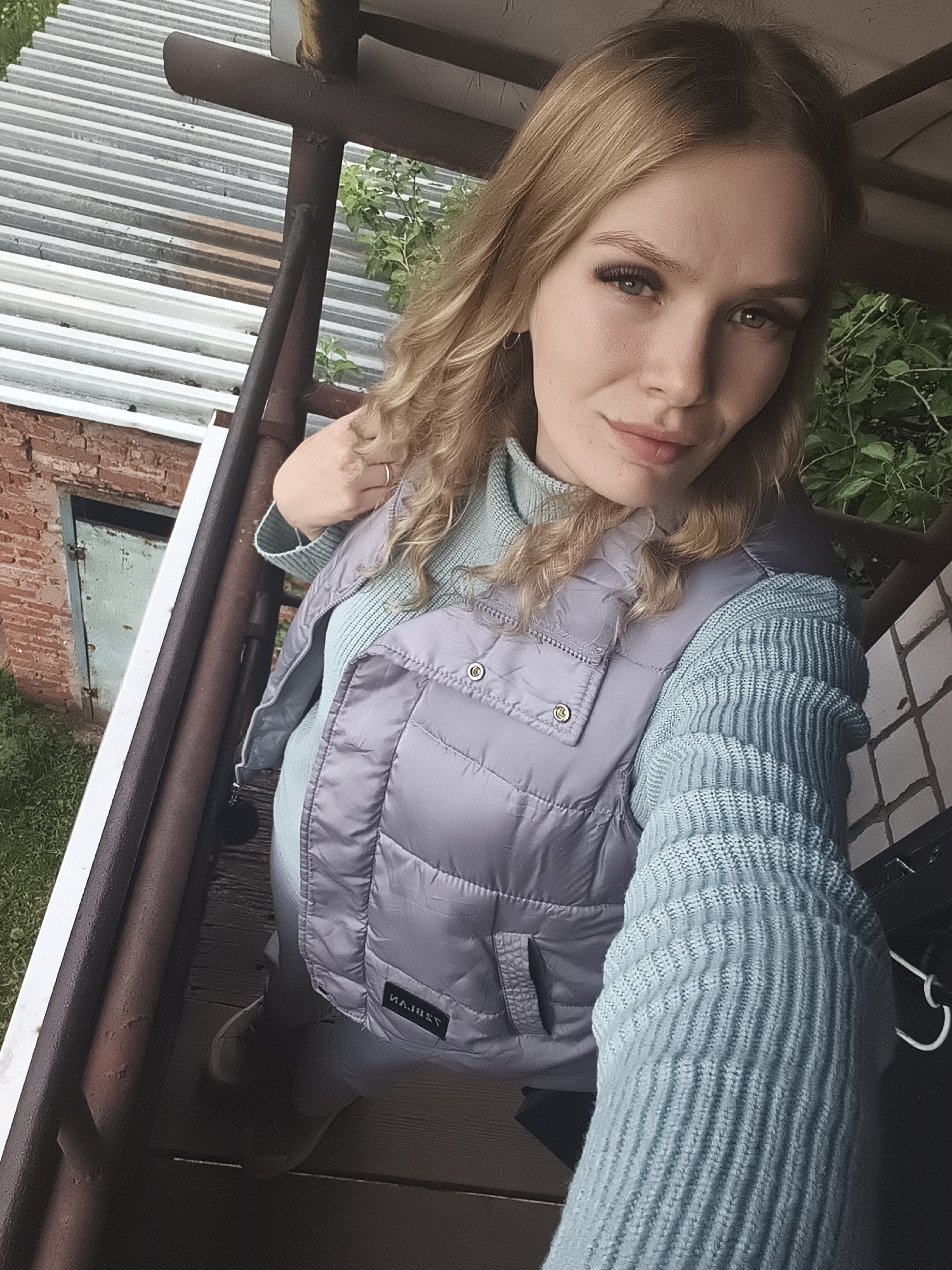 A young woman with long, wavy blonde hair and light-colored eyes is captured in a selfie. She wears a light blue ribbed knit turtleneck sweater beneath a light grey quilted puffer vest. Her right hand, wearing a ring, is positioned near her neck, while her left arm extends, likely holding the camera. She looks directly at the viewer with a composed expression.
The setting appears to be an elevated outdoor structure, possibly a balcony or porch, with rusty brown metal railings and supports around her. Below her feet are wooden planks. In the background to her left, a red brick building with a faded light blue door and green grass is visible, topped by a corrugated metal roof. Green foliage and trees are present in the background to her right. The lighting suggests it is daytime, with soft, even light, possibly indicating an overcast sky. A label on the pocket of her vest shows the reversed text "MAJESS". The scene takes place in Русский Вожой, Russia.