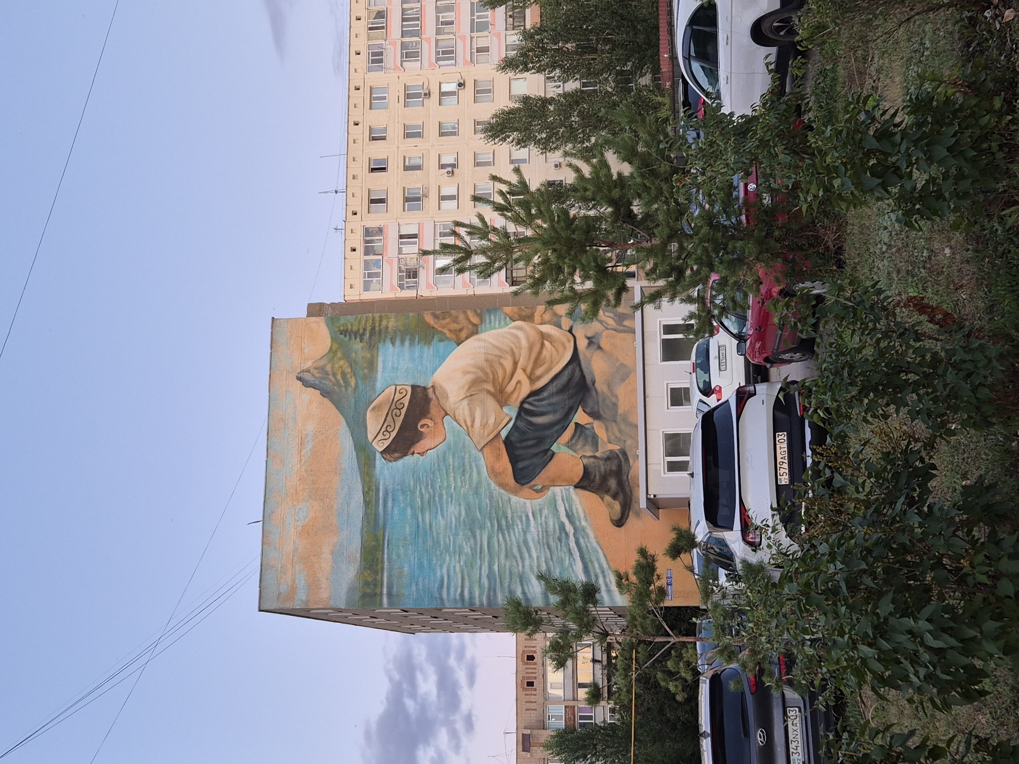 The image depicts a large mural painted on the side of a multi-story apartment building in Kokshetau, Kazakhstan. The mural features a young boy wearing a traditional Kazakh hat, sitting on a rocky shore by a body of water, with mountains in the background under a pale blue sky with some clouds. The apartment building adjacent to the mural is a light-colored, multi-story structure with numerous windows and balconies. In the foreground, parked cars are visible, partially obscured by green bushes and small coniferous trees. The scene appears to be an outdoor urban environment, likely a residential area.