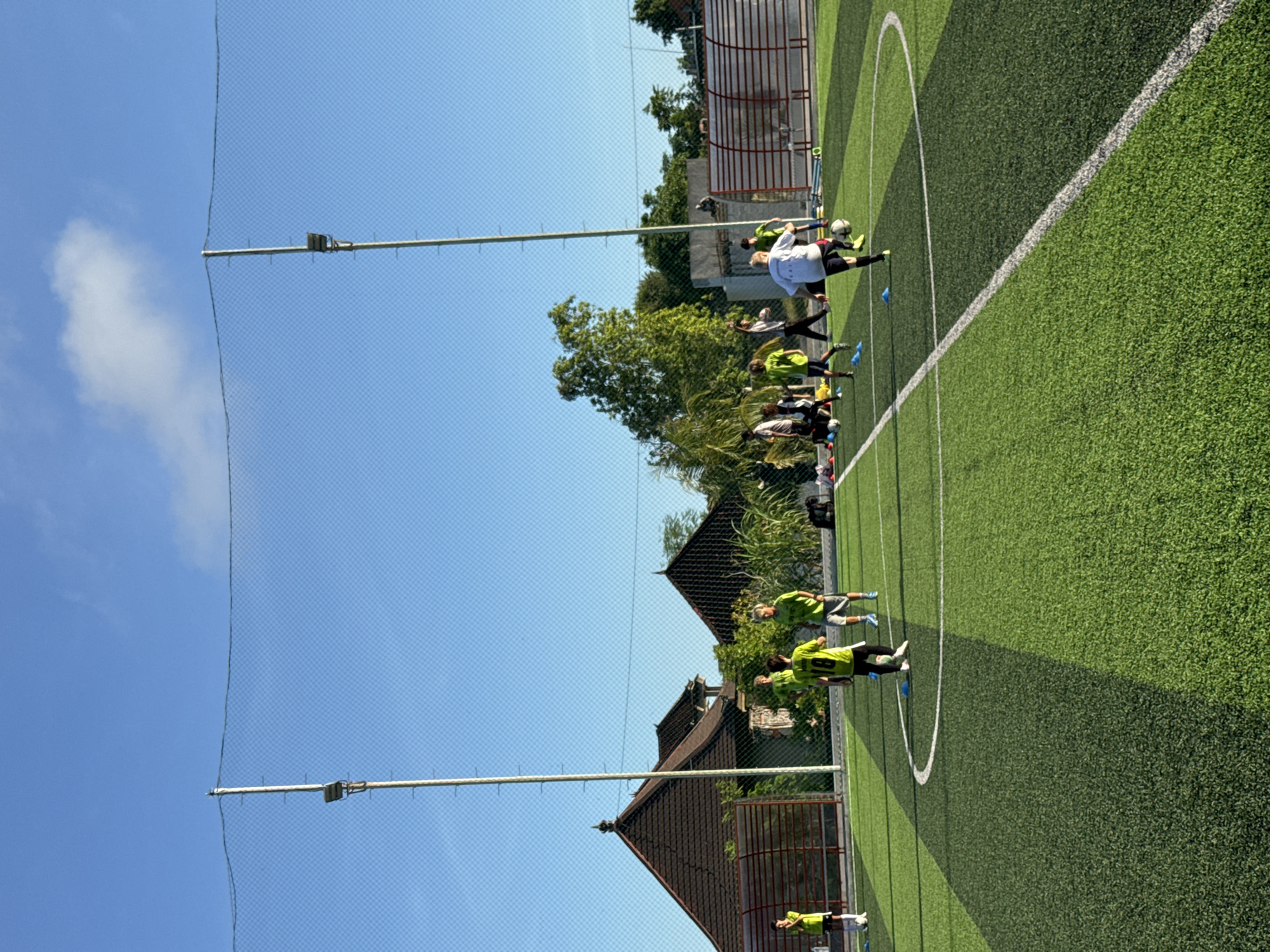 A soccer practice is underway on a green field in Kuta Selatan, Indonesia. A group of soccer players are practicing in the field. The sky is a bright blue with light white clouds. A net is in the foreground, reaching up to the sky. Behind the field, there are buildings and trees.