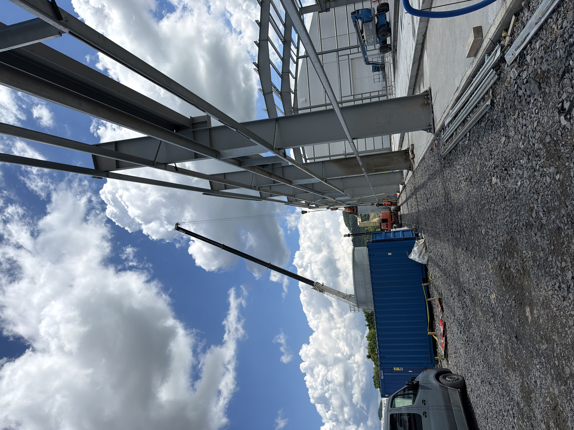 A construction site is depicted in Stróża, Poland, under a partially cloudy blue sky. In the foreground, a grey van is parked on a gravel surface, to the left of a large blue shipping container. Several pieces of metal scaffolding and a white bag are also on the ground near the container. The main focus of the image is the skeletal structure of a large building under construction, composed of grey steel beams. A tall mobile crane with a black boom is positioned behind the shipping container, with its arm extended upwards. Further in the background, an orange construction vehicle is visible, as well as a blue aerial work platform. A lighter-colored, cylindrical structure with a flat top stands behind the blue container. The environment suggests an active construction project with materials and machinery present.
