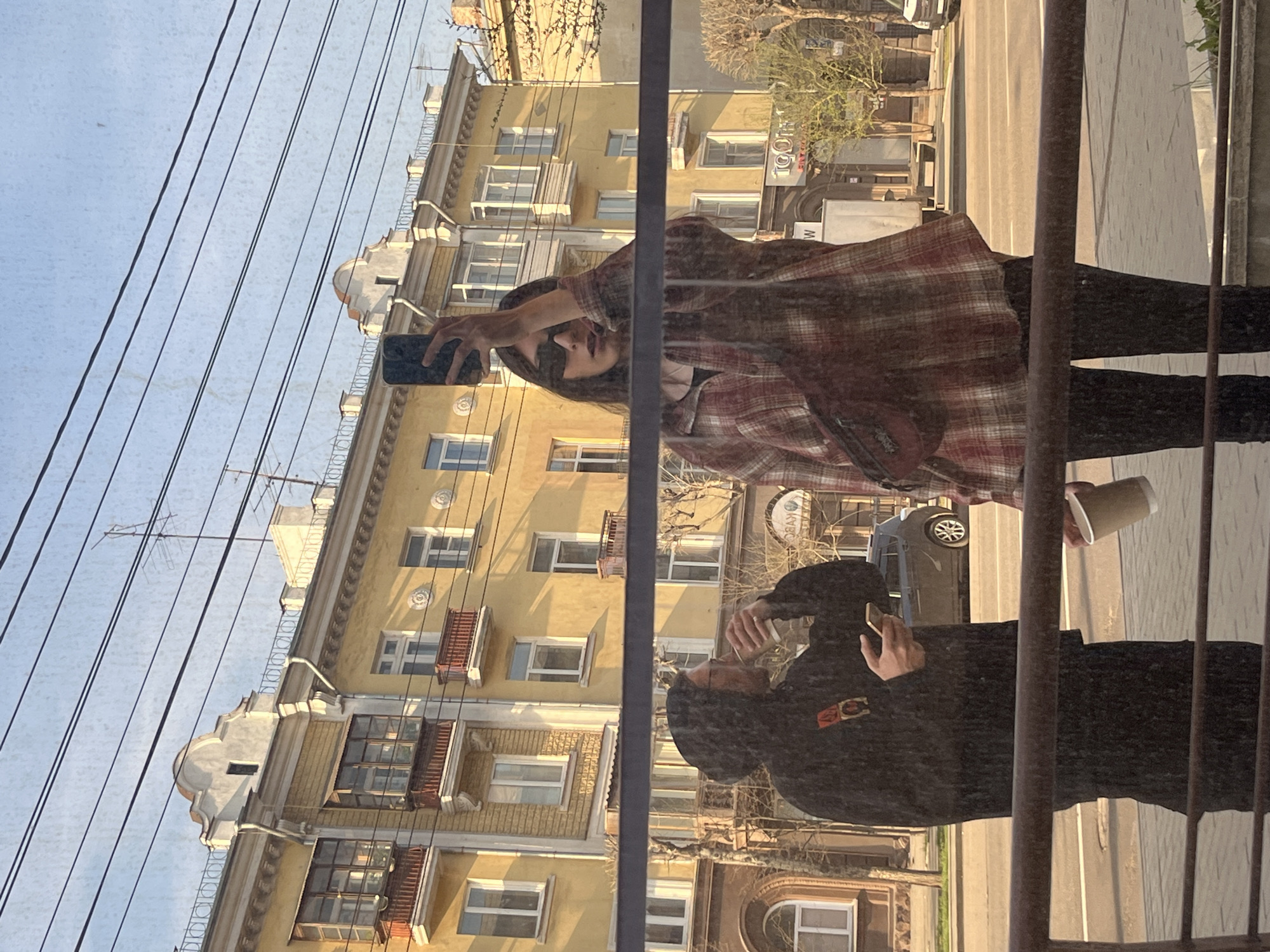 A vertically oriented, eye-level shot captures a reflection in a window. The reflection includes two figures, a yellow building, and a sky with overhead wires. A woman in a plaid shirt holds a phone up as if taking a selfie. She has dark hair and is wearing sunglasses. A man in a dark hooded jacket appears to be holding a phone to his ear and carrying a cup. The building features multiple windows and decorative architectural details. The environment outside appears to be a city street. The window also reflects the street with a car, trees, and utility poles. A vertical set of bars is placed in front of the window, possibly part of a railing or fence.
Based on the "KYBC" sign visible in the reflection, the location is near the Kryvbas Restaurant on Mira Avenue in Krasnoyarsk, Russia.