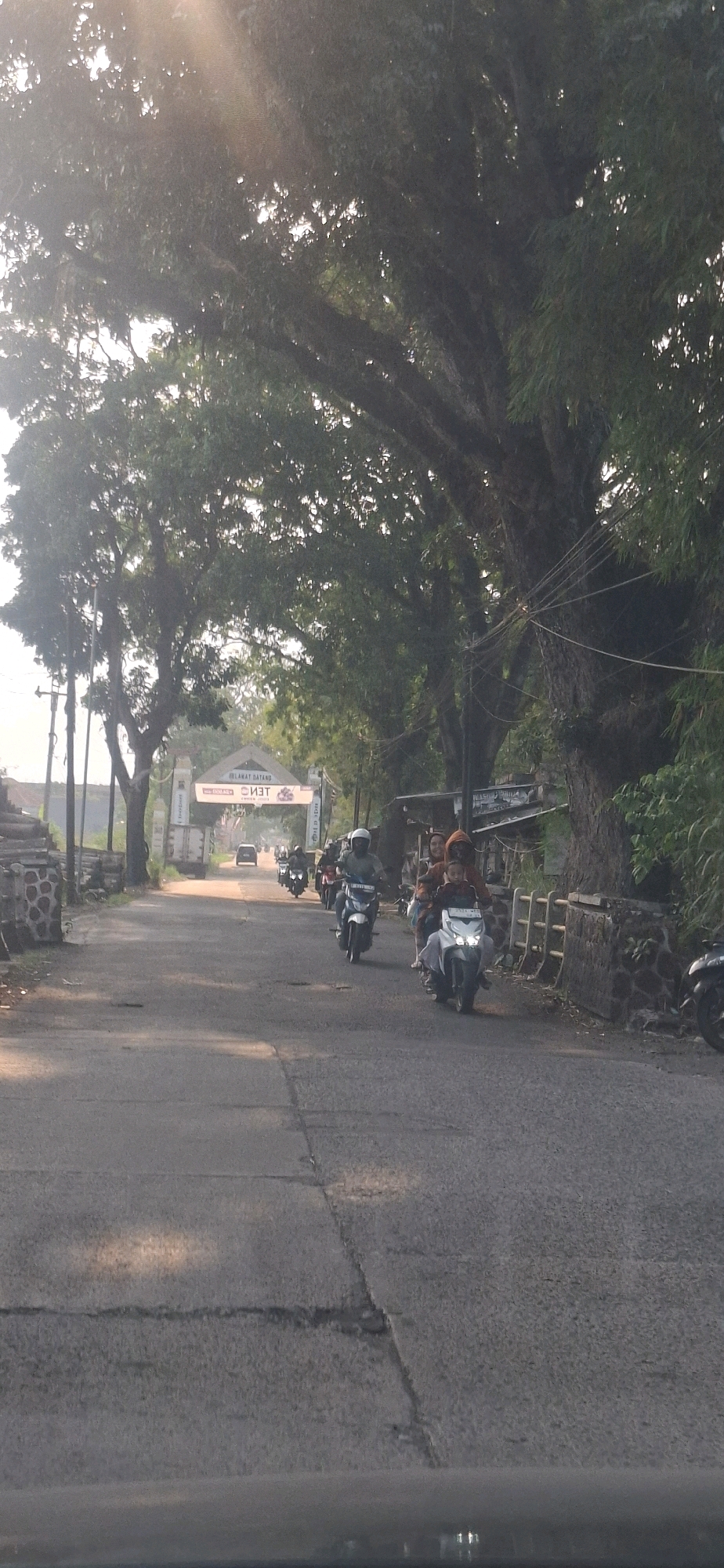 The image captures a sun-drenched road in Sukasari, Indonesia, lined with large, mature trees whose branches create a dappled canopy overhead. A few motorcycles are seen traveling along the asphalt road. One motorcycle in the foreground carries three individuals, two adults and a child, all looking forward. Another motorcycle, slightly ahead and to the left, has a single rider wearing a helmet. In the distance, a white car is visible, and further down the road, an archway with text that reads "SELAMAT DATANG" (Welcome) is illuminated. The environment suggests a somewhat rural or semi-urban setting with a sign indicating an entrance. The lighting indicates it is daytime, with sunlight filtering through the leaves.