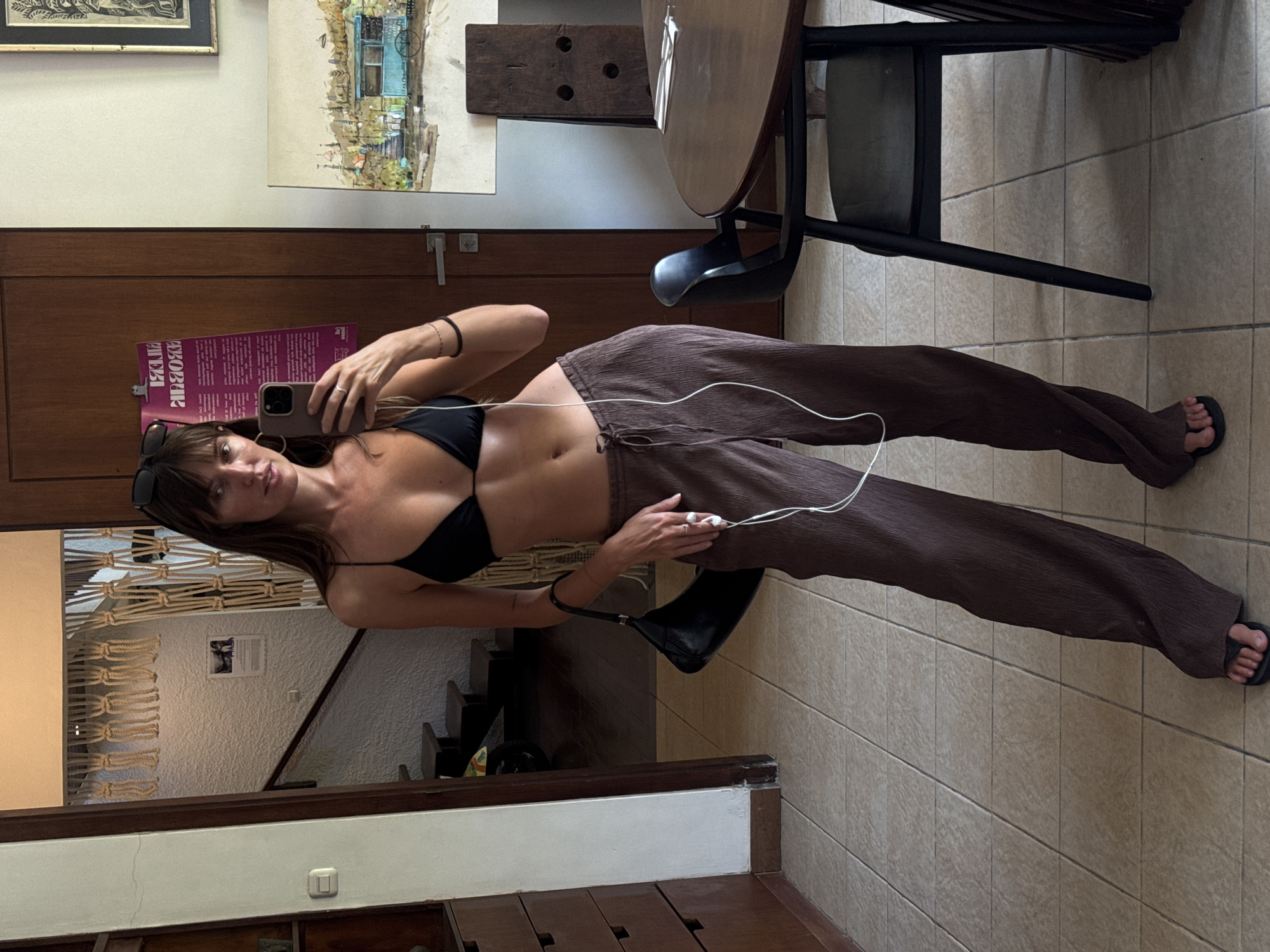 A woman stands in a room, taking a selfie in a mirror. She is wearing a black bikini top and brown, pleated, wide-leg pants. She holds a smartphone in her right hand and has earbuds in her ears. A black handbag hangs from her left shoulder. The room appears to be a home with tiled floors and wooden furniture, including a round table and chairs. In the background, a staircase leads to another level, and macrame decorations hang on the wall. The overall impression is of a casual, indoor setting.