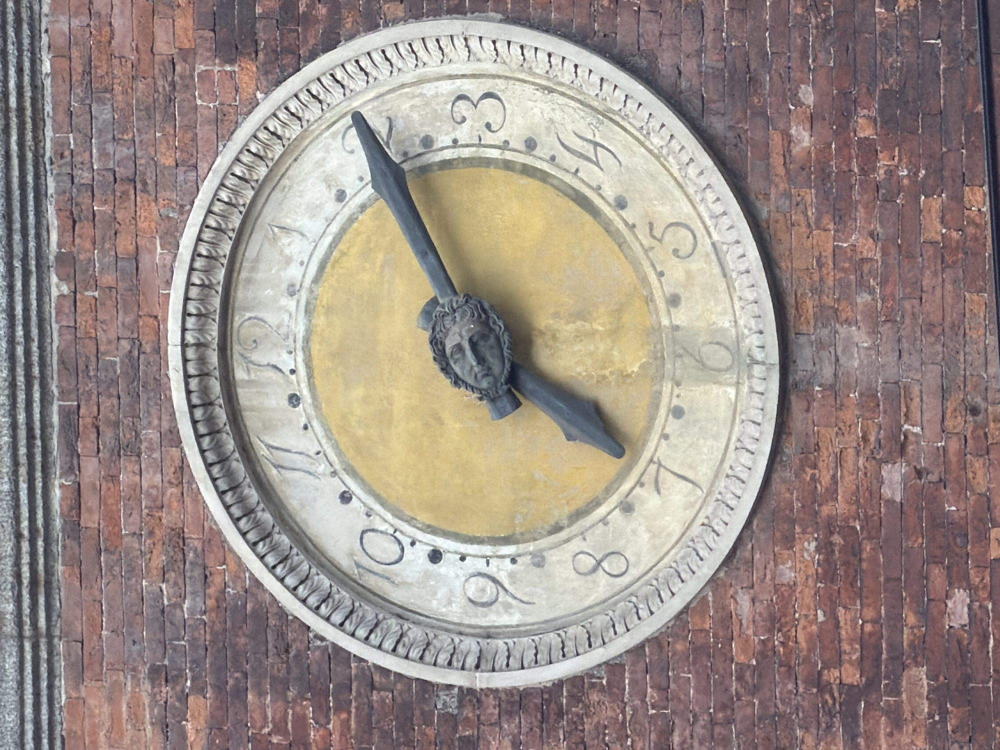 A detailed, eye-level shot captures an ancient, ornate clock face mounted on a weathered red-brick wall, typical of historic architecture in Milan, Italy. To the far left, a section of a fluted grey stone column or pilaster is visible, suggesting the clock is part of a larger building facade.
The clock face is circular, with an off-white or light grey outer ring featuring intricate carved patterns, resembling a laurel wreath or similar decorative motif. The central part of the clock face is a warm golden yellow. Around the perimeter of the golden center, highly stylized black numbers or symbols mark the hours. Clearly discernible are a '3', a stylized '4' (resembling an 'h'), a '5', and an '8' (depicted as an infinity symbol, '∞'). Small black dots are interspersed between some of these markings.
The clock has two dark, metallic hands. The shorter, hour hand is embellished with a detailed, classical-style sculpted face, likely male, positioned at its base where it attaches to the clock mechanism. This hand points towards the '3'. The longer, minute hand is simpler in design and points towards the '9' or '10' position (around the 45-minute mark). Thus, the clock indicates approximately 3:45.
The brick wall behind the clock shows signs of age, with varying shades of reddish-brown and some mortar deterioration, adding to the historic character of the scene. The overall lighting is soft and even, suggesting either an overcast day or indirect light, highlighting the textures and details without harsh shadows. No human activity is depicted; the image focuses solely on the architectural and timekeeping elements.