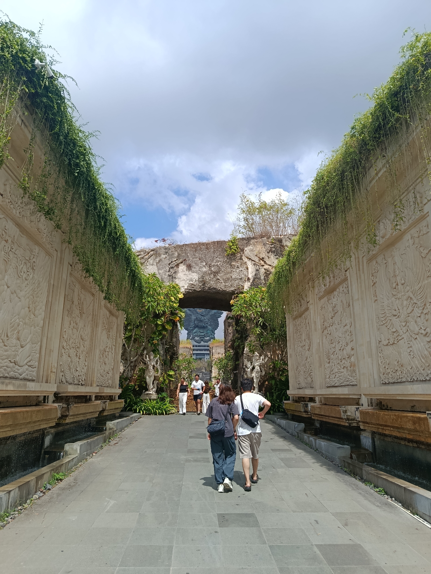 In Kuta Selatan, Indonesia, a pathway paved with large gray tiles leads towards an ancient-looking stone archway. On either side of the pathway are walls adorned with intricate, light-colored bas-relief carvings depicting mythical figures and scenes. Lush green vines cascade down from the tops of these walls, adding a touch of nature to the architecture. In the foreground, a couple walks away from the camera, the woman in a dark top and blue pants, the man in a white t-shirt and light brown shorts. Other pedestrians can be seen in the distance, walking towards the archway. Beyond the archway, a large, dark statue of what appears to be a mythical creature is visible, with a building complex behind it. The sky above is a mix of blue with scattered white clouds. The scene suggests a cultural or historical site, possibly a temple complex or a heritage area.