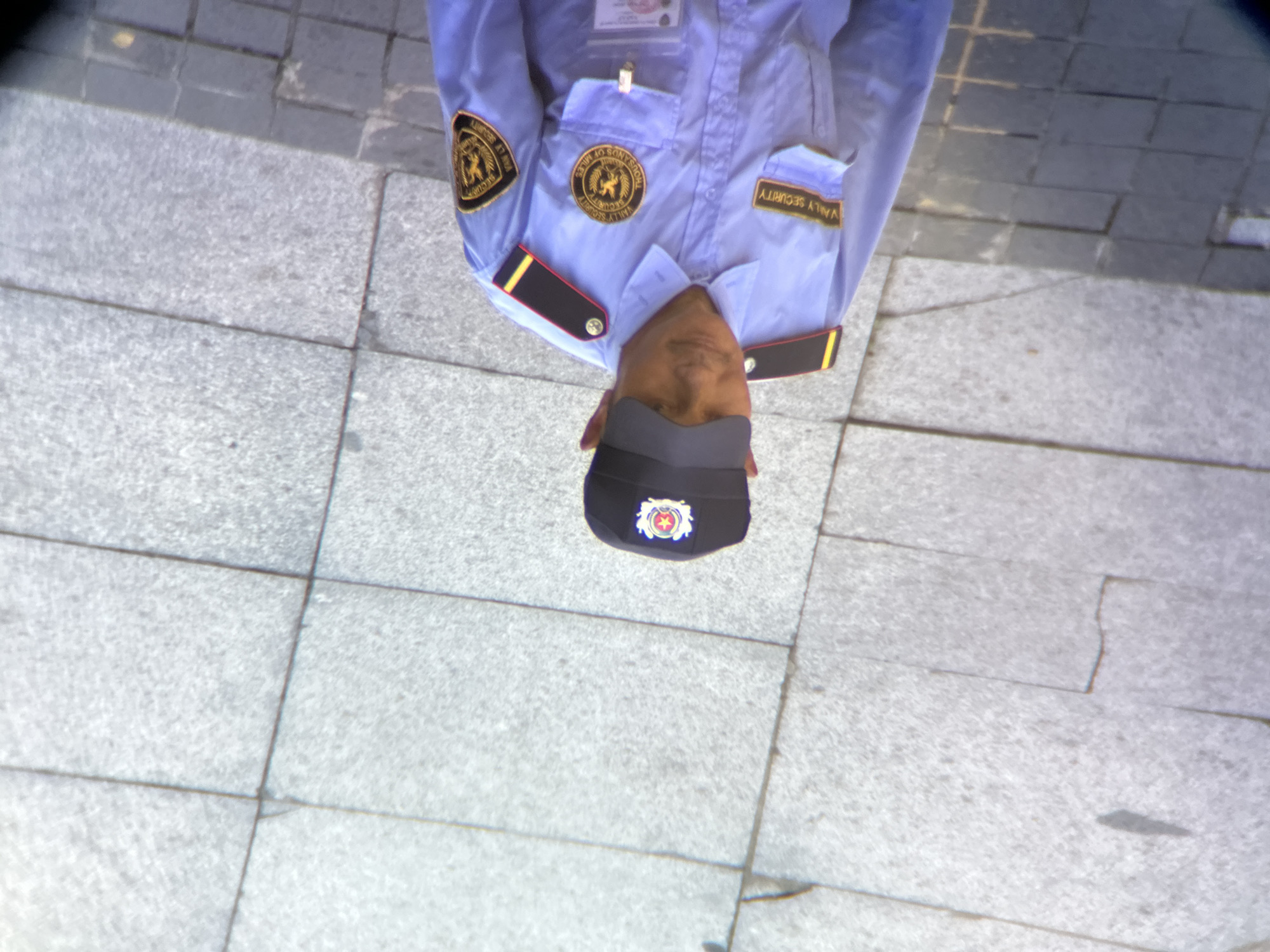 A security guard in a light blue uniform stands on a paved walkway in Đà Nẵng, Vietnam. The guard wears a black cap with a red and gold insignia, and a blue shirt with patches on the shoulders and chest. One patch on the left chest reads "VALLEY SECURITY", and another circular patch on the right chest depicts a lion and text. A clear ID badge is clipped to the front of the shirt. The ground is made of large, grey stone tiles with visible grout lines.
