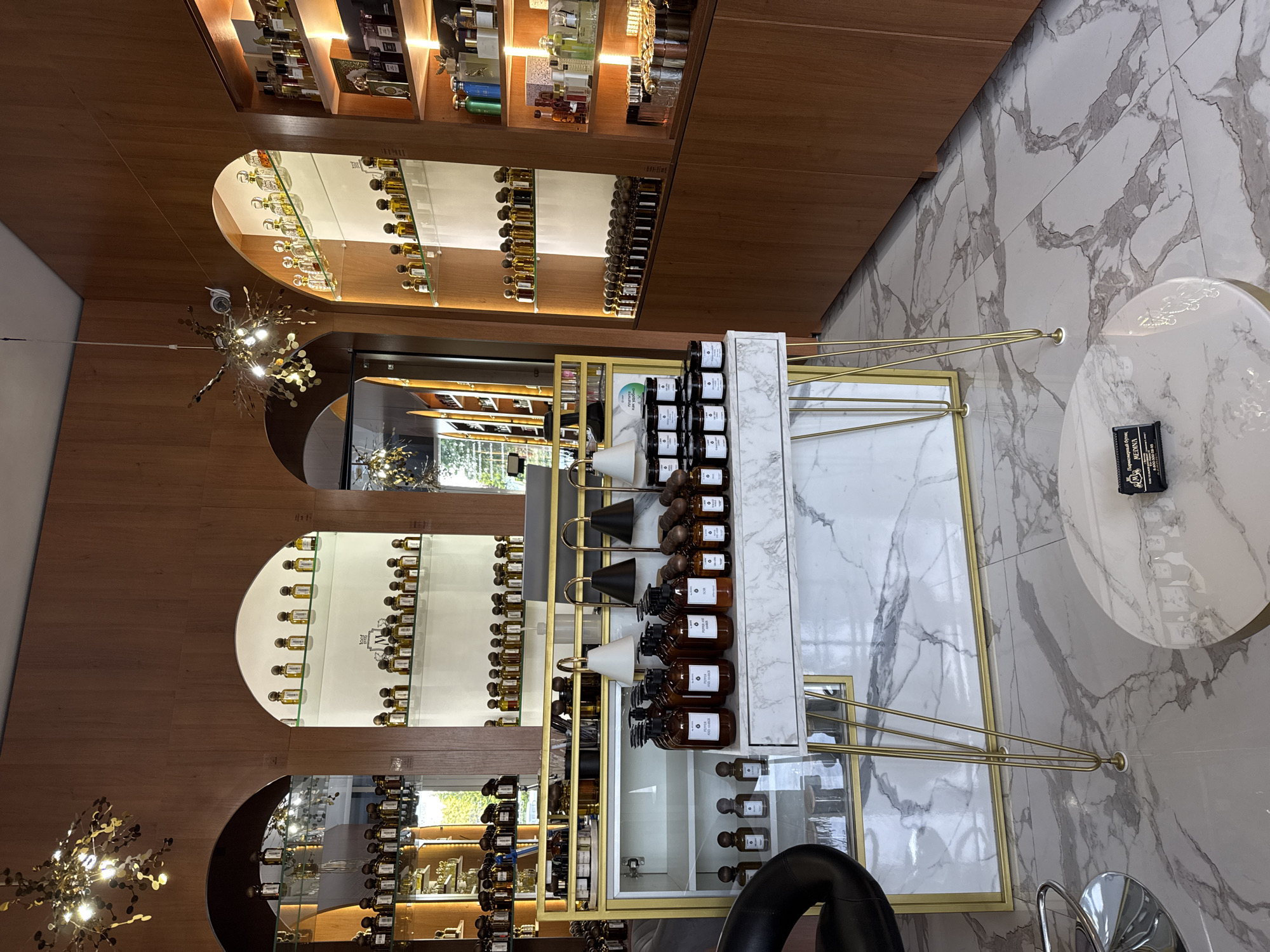 The image shows the interior of a perfume store, likely in Krasnodar, Russia, given the text on the business card on the table in the foreground. The store features arched displays with numerous bottles of perfume and other fragrance products. The walls are paneled with warm-toned wood, and the lighting is soft and inviting. In the foreground, a white marble-topped display table with gold hairpin legs holds several dark-colored perfume bottles. Behind this table, a partially visible black leather chair is present. To the right, tall wooden shelving units are filled with a variety of perfume bottles and boxes. The floor is tiled with a marble-effect pattern. The overall impression is one of a luxurious and well-organized retail space.