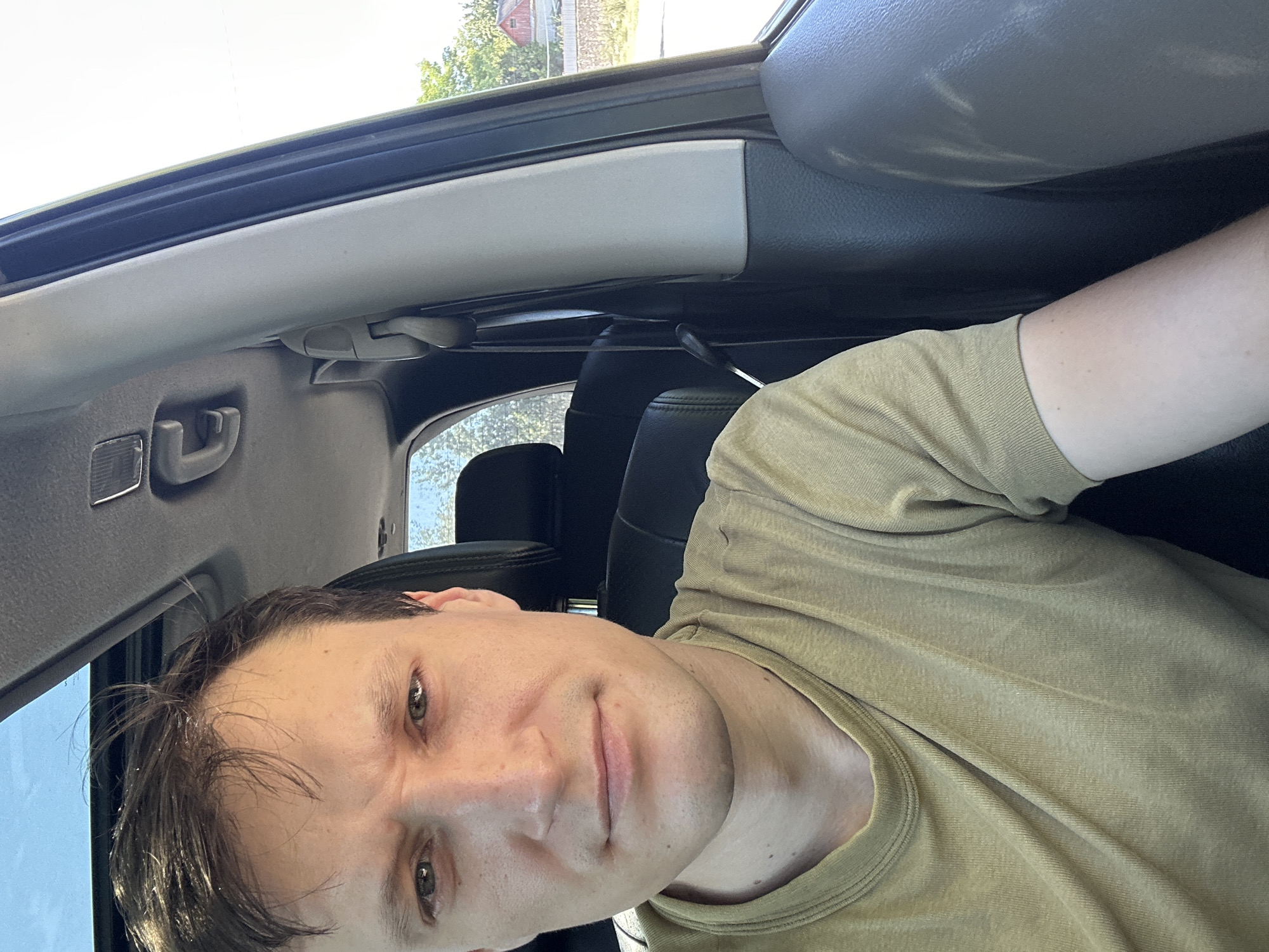 A man with short brown hair and fair skin is sitting in the driver's seat of a car, taking a selfie. He is wearing a plain olive-green t-shirt. His face is directed towards the camera, with his eyes looking slightly to the left. The interior of the car is visible, including the headrest of the passenger seat behind him and part of the car's sunroof. The sun is shining brightly, casting shadows within the car. Outside the car window, trees and a building with a red roof can be seen. The image appears to have been taken during the daytime.