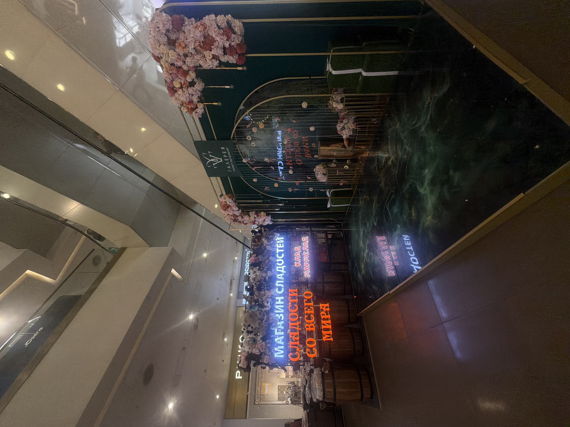 The image shows an indoor shopping mall environment. In the foreground, there is a dark, glossy floor that reflects the lights and signage above. To the left, there are several wooden barrels, some of which appear to be covered with fabric. Behind the barrels, a prominent neon sign reads "МАГАЗИН СЛАДОСТЕЙ" (Candy Store) in blue letters, with "СЛАДОСТИ СО ВСЕГО МИРА" (Sweets from all over the world) in orange letters below it. A smaller sign also reads "КЛАД МАРМЕЛАД" (Marmalade Treasure). A collection of stuffed animals is piled above the neon signage.
To the right of the candy store display, there is a decorated backdrop with a dark green wall and gold archways. This backdrop is adorned with floral arrangements in shades of pink and white, and hanging strings of small lights and decorative elements. A logo on a green banner at the top of the backdrop reads "ГАЛЕРЕЯ САНКТ-ПЕТЕРБУРГ" (Gallery Saint Petersburg). There are also gold pedestals and green moss-covered boxes with white ribbons, resembling gifts, placed in front of the backdrop.
The overall impression is a festive or promotional display within a commercial setting. The lighting suggests it is either evening or the mall's interior lighting is predominantly artificial. The presence of the "Gallery Saint Petersburg" logo indicates the location is likely a shopping gallery or mall in Saint Petersburg, Russia. There are no people visible in the image.