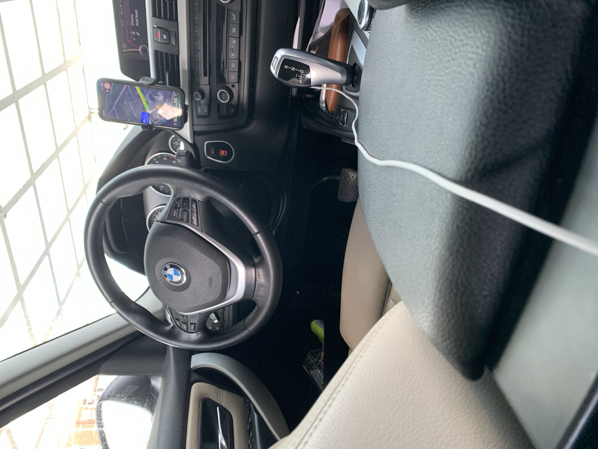 The image displays the interior of a car, specifically the driver's perspective looking towards the front and passenger side. The car appears to be a modern BMW model, indicated by the steering wheel emblem and dashboard design.
In the foreground, a black leather steering wheel with a silver trim and a BMW logo at its center is prominent. Text "AIRBAG" is visible on the steering wheel. To the left of the steering wheel, through the driver's side window, bright light suggests it is daytime, and a patterned structure, possibly a building or garage window, is reflected.
Mounted to the right of the steering wheel, on the dashboard, is a smartphone displaying a map or navigation application. Text "14:53", "iPhone", and "AUX front" are visible on its screen. Below the smartphone, the central dashboard features climate controls, a radio unit, and various buttons, including a hazard light button and a button with "OFF" text.
Further to the right, a silver-finished automatic gear shifter is visible, showing "P", "R", "N", "D", and "M/S +/-" settings. A white charging cable extends from the center console area, across the gear shifter, towards the passenger side. The passenger seat is upholstered in light beige or cream-colored leather. On the floor mat on the passenger side, a neon green object and a clear container are partially visible.
The overall scene suggests the car is stationary, possibly parked indoors or in a garage due to the bright, uniform light and reflections. No people are visible.