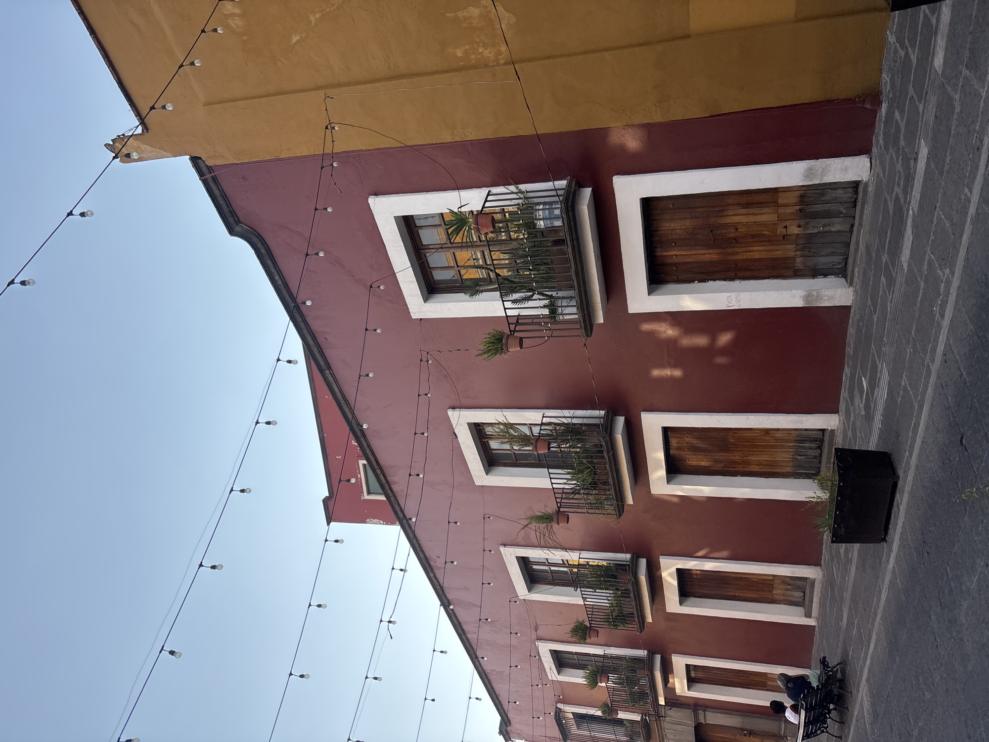 The image depicts the architecture of a building in Puebla, Mexico. The building is a burgundy/red color with a golden-yellow roof, and multiple windows with white frames. Some windows have plants, and some are boarded up with wood. A string of lights is running along the facade of the building and across the sky. The building abuts a paved street area with a potted plant. Two figures are visible near the bottom right of the image, seated on a bench.