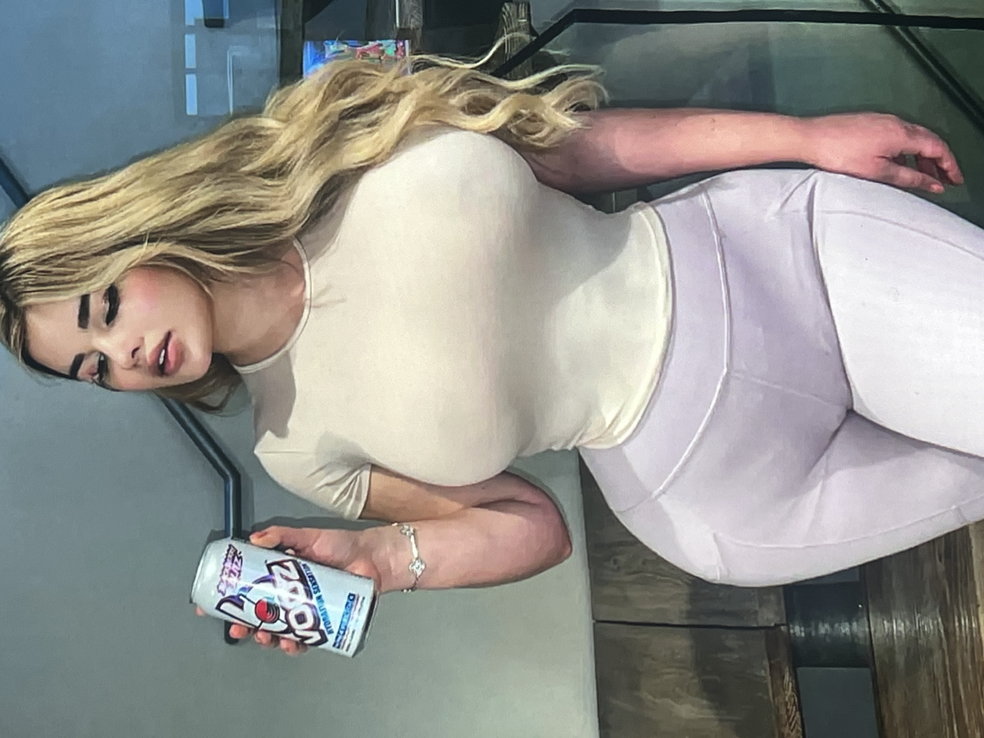 A woman with blonde hair is holding a can of "VOYZ Hydration Sensation" drink. She is wearing a cream-colored short-sleeved top and light purple leggings. The background appears to be an indoor setting with wooden benches and a railing. There is no indication of the city of Vanadzor, Armenia in the image.