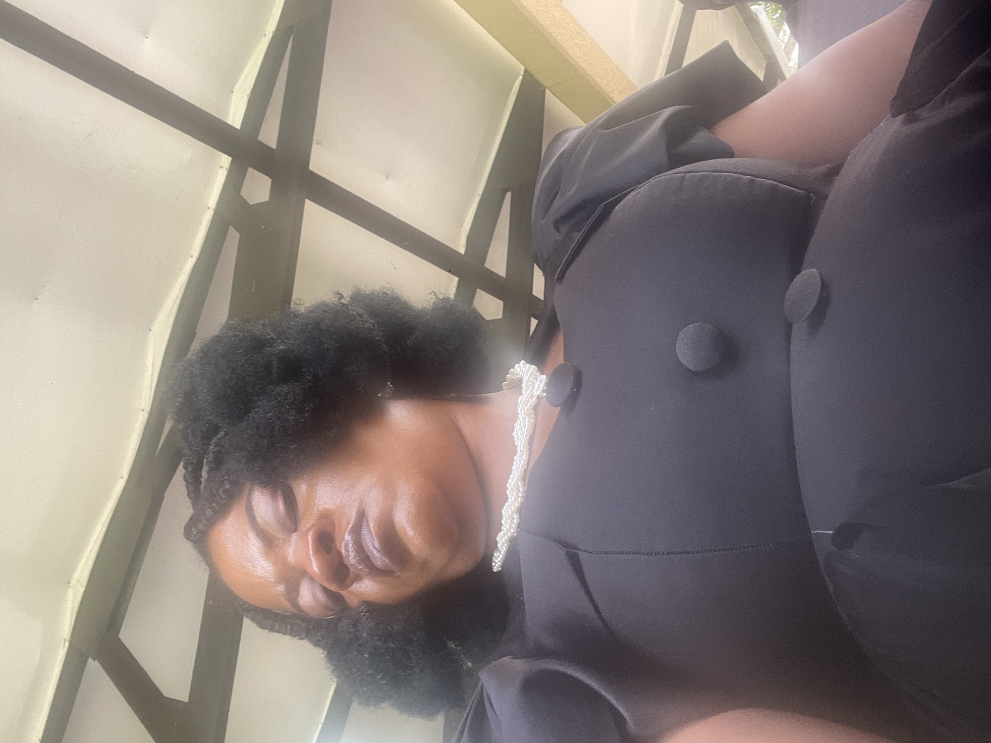 The image shows a woman in a black dress and pearl necklace, seemingly asleep or resting. She is positioned low in the frame, with her head tilted back and eyes closed. The background reveals a light-colored ceiling or canopy with dark structural beams, suggesting an outdoor sheltered area. The overall lighting is bright, indicating daytime. The scene is devoid of any specific landmarks that would place it in Itoga, Nigeria.