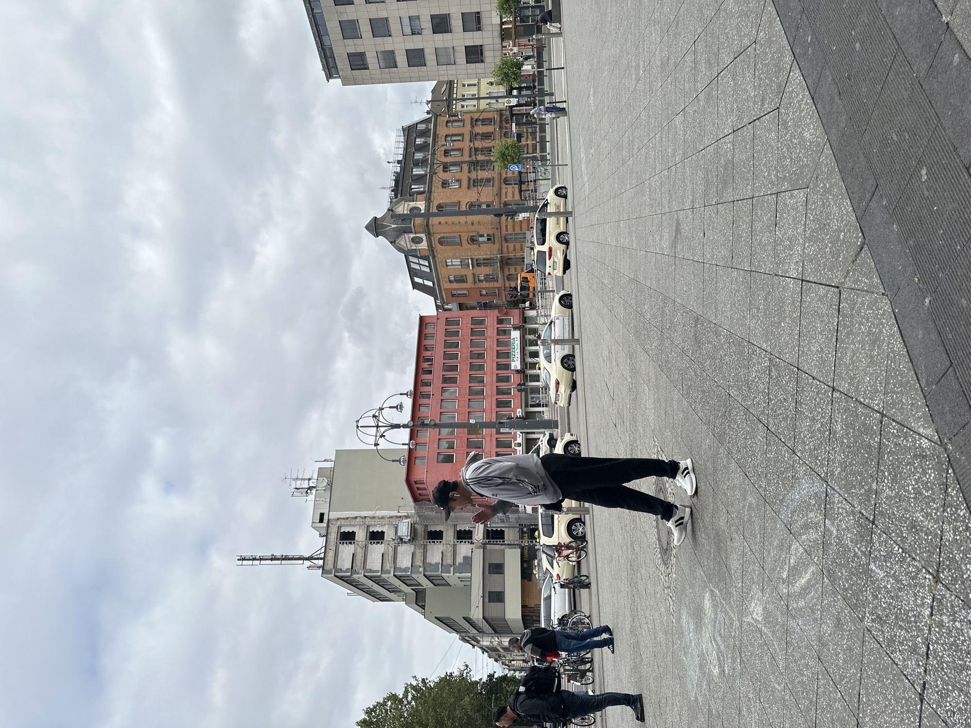 The image depicts a street scene in Krefeld, Germany, likely a public square or intersection. In the foreground, a man wearing a grey tracksuit and a black baseball cap is standing on a paved surface, appearing to gesture with his right hand. His left hand is in his pocket. He is facing towards the right of the frame. Behind him, to his left, another man in a dark jacket and dark pants is visible, also standing. Further in the background, several people are walking or standing near bicycles.
The environment includes various buildings: a tall, partially demolished or under-construction building with visible scaffolding on the left, a reddish-pink multi-story building with many windows in the middle ground, and an older, ornate brick building with a distinctive roofline to its right. Another modern, grey building is on the far right. Several yellow taxis are present on the street, one prominently in the middle ground. The sky is overcast with a cloudy, grey appearance. The ground is paved with large square tiles, some of which appear to have chalk markings or dirt. Streetlights and traffic signals are also visible. The overall impression is a bustling urban environment on a cloudy day.