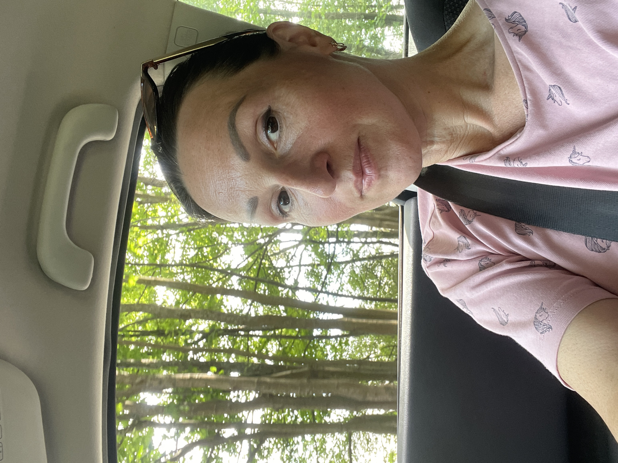 A woman with dark hair is taking a selfie in the back seat of a car. She is wearing a pink t-shirt with a unicorn pattern and a seatbelt is visible. Sunglasses are perched on top of her head. The background shows trees and foliage seen through the car window, suggesting a natural setting outside the city of Zelenogorsk, Russia. The interior of the car, including the headrest and a handle, is also visible.