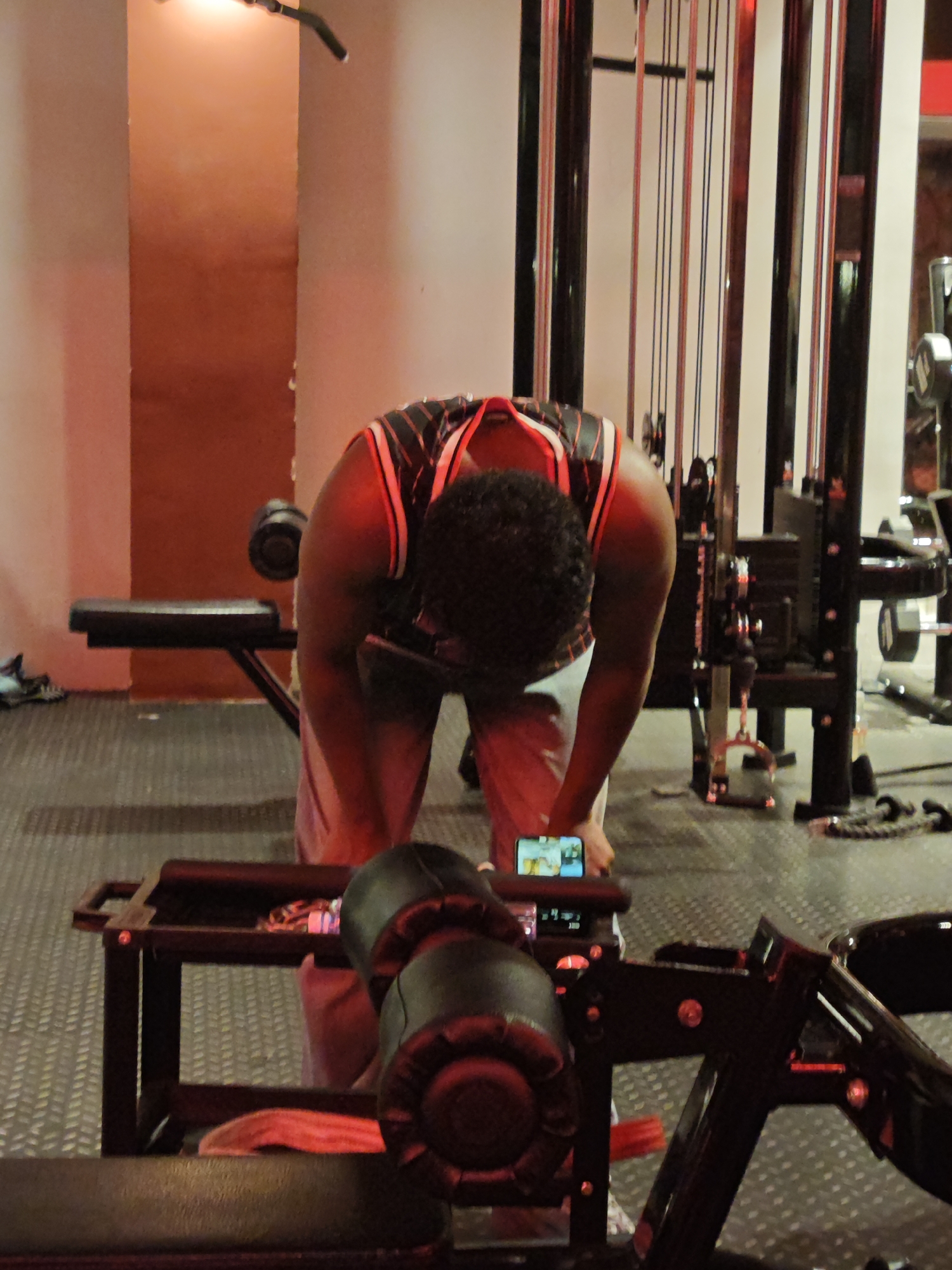 The image, possibly taken in a gym within the Chongqing High-tech Zone in China, shows an individual bent over with a phone in their hand. The person is wearing a tank top with black and red trim, and light-colored pants. They are standing near gym equipment. A phone is set up to view the person. The surrounding environment includes gym equipment, a black bench, and flooring with a pattern. The room is illuminated with a mix of natural and artificial light.