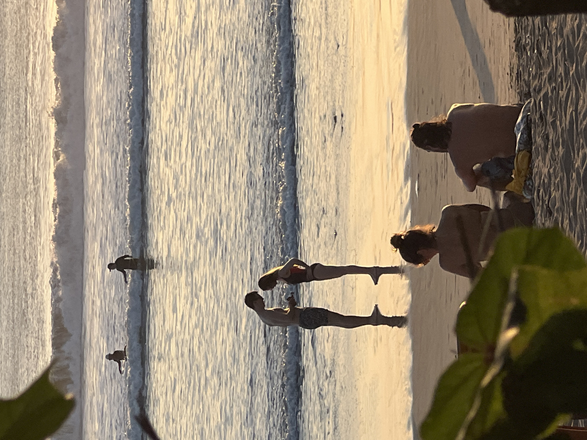 In Kuta, Indonesia, at sunset, a couple stands near the water's edge on a beach, silhouetted against the warm light. The man, wearing patterned shorts, faces the woman, who is in a dark swimsuit. They appear to be looking at something in the man's hand. In the background, two other figures are visible in the ocean, one on a surfboard and another standing in the water. In the foreground, two people sit on the sand with their backs to the camera, facing the ocean. Lush green foliage partially obscures the left side of the frame. The ocean displays gentle waves, reflecting the golden hues of the setting sun. The wet sand near the water's edge creates a mirror-like effect.