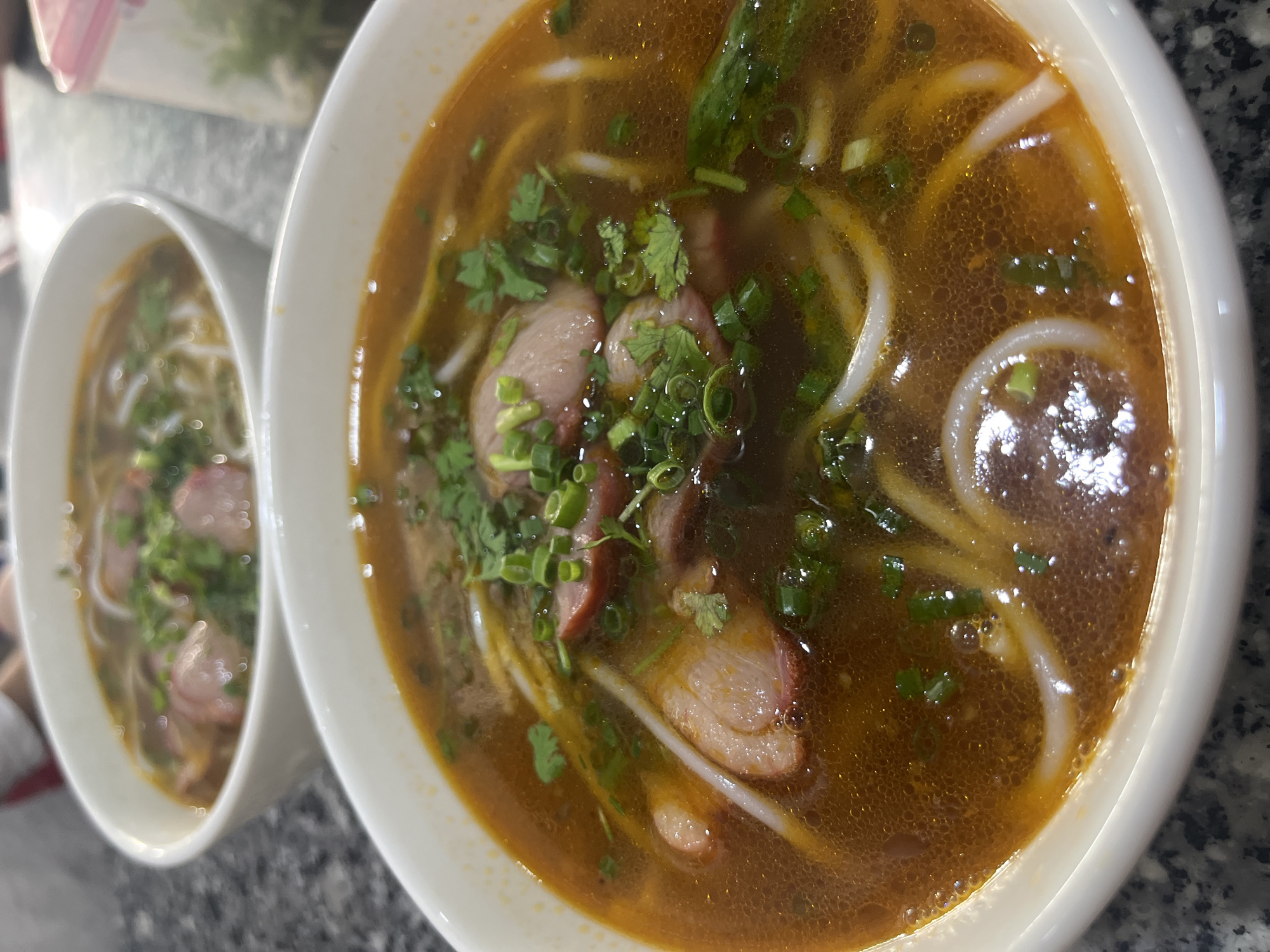 A close-up, eye-level shot focuses on two bowls of Vietnamese noodle soup, known as "Bun Bo Hue", on a speckled gray countertop. The bowl in the foreground is larger and more in focus, filled with a rich, reddish-brown broth. Slices of red meat, likely beef or pork, are visible submerged in the broth, along with thick white noodles and chopped green onions and cilantro as garnish. Bubbles are visible on the surface of the broth, indicating it is hot. The second bowl, positioned behind and to the left of the first, is slightly out of focus but appears to contain a similar noodle soup. To the right of the bowls, a clear plastic container with a pink lid is partially visible, containing chopped green herbs. The lighting is natural and diffused. The overall impression is a casual, home-style meal in Vietnam, likely in the Bac Nha Trang Ward.