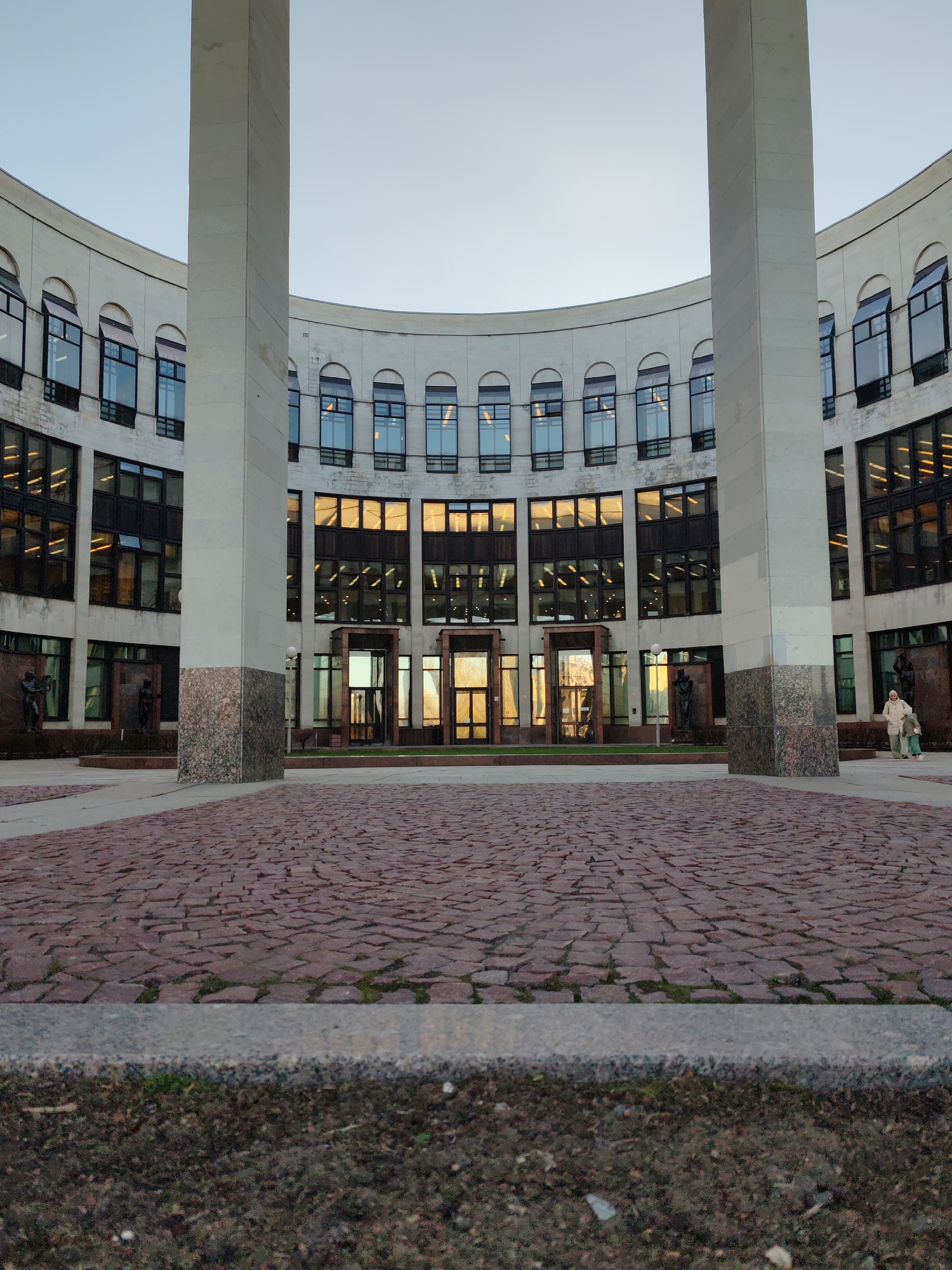 The image shows the entrance to the Saint Petersburg State University of Economics in Saint Petersburg, Russia. The curved building facade is made of white stone with arched windows on the upper level and rectangular windows on the lower level. Two large square pillars flank the entrance doors. The ground is paved with red bricks and there is a patch of dirt in the foreground. Two people are standing near the right side of the building. There are bronze statues in front of the building. The sky is overcast.