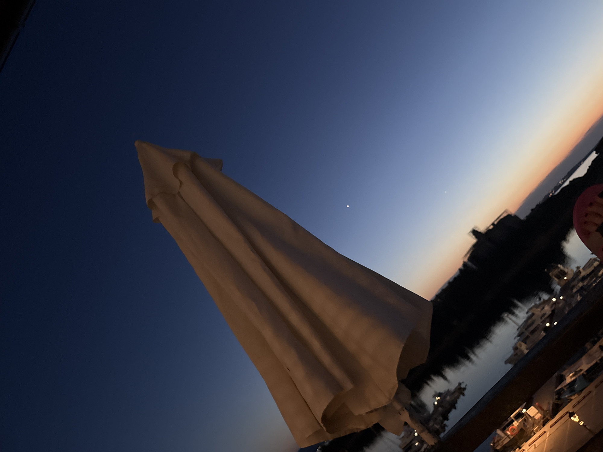 The image captures a dusk scene in Sozopol, Bulgaria, with a closed, light-colored umbrella prominently featured in the foreground, angled upwards against a darkening sky. The sky transitions from a deep blue at the top to a soft orange and pink gradient near the horizon, indicating the twilight hours. A single bright star or planet is visible in the upper portion of the sky.
In the background, a body of water is visible, likely the sea, with reflections of buildings and lights. These lights suggest a harbor or a waterfront area with boats and structures lining the shore. The silhouettes of buildings are dark against the twilight sky, hinting at a developed coastal town. The overall mood is peaceful and serene, typical of a sunset or dusk by the water. There are no visible people in focus, though a hint of a person's foot wearing a pink sandal is visible on the far right edge of the frame. No text is present in the image. The weather appears calm, with no indication of wind or rain.