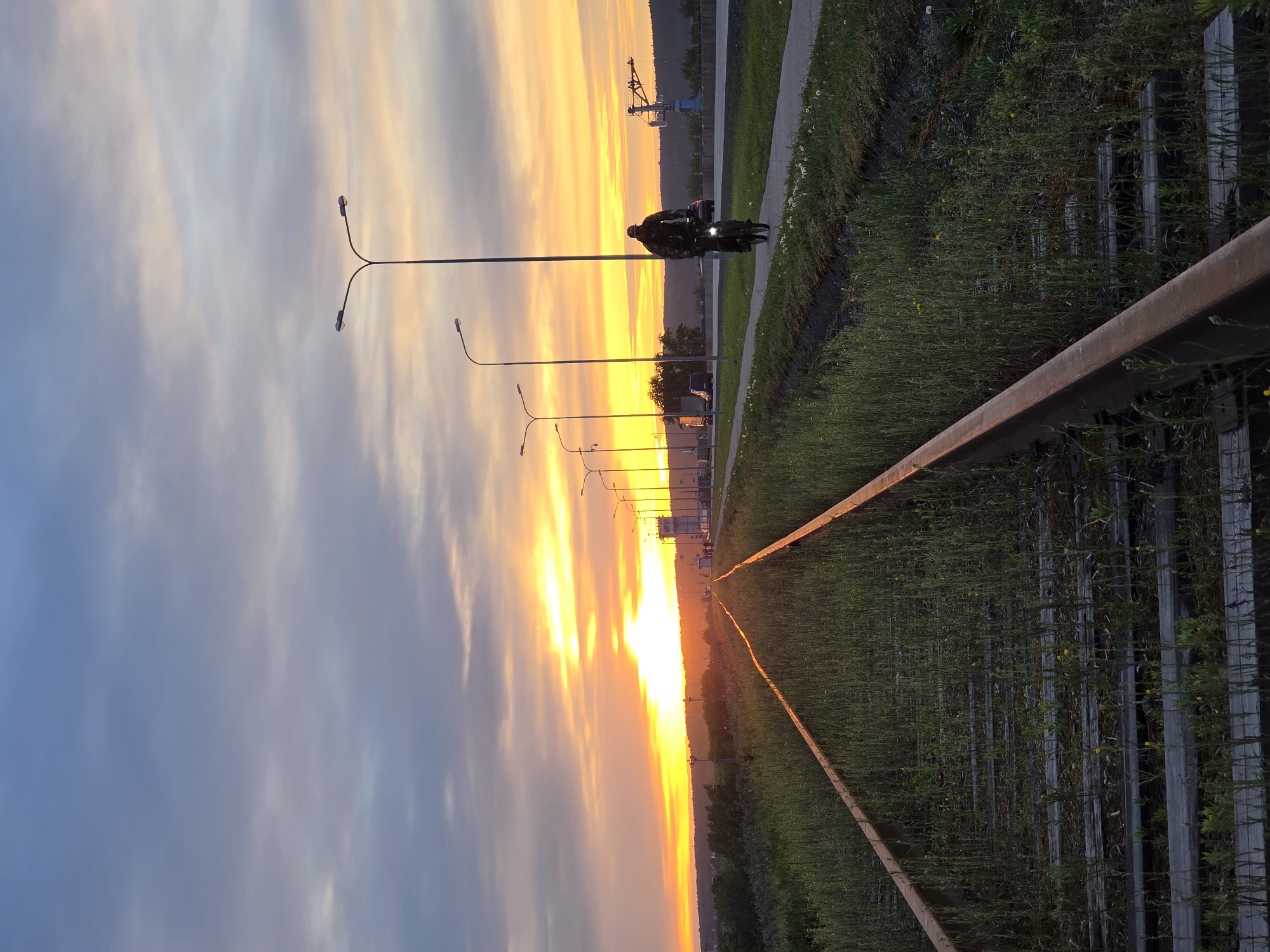 The image shows a motorcycle with two people on it riding on a sidewalk in Chaikovsky, Russia. The image is taken at sunset, with the sun setting in the distance. The sky is orange and yellow, and the clouds are gray and blue. There are streetlights along the sidewalk. A fence or railing runs along the right side of the image, next to a grassy area. A crane is visible in the background on the horizon.