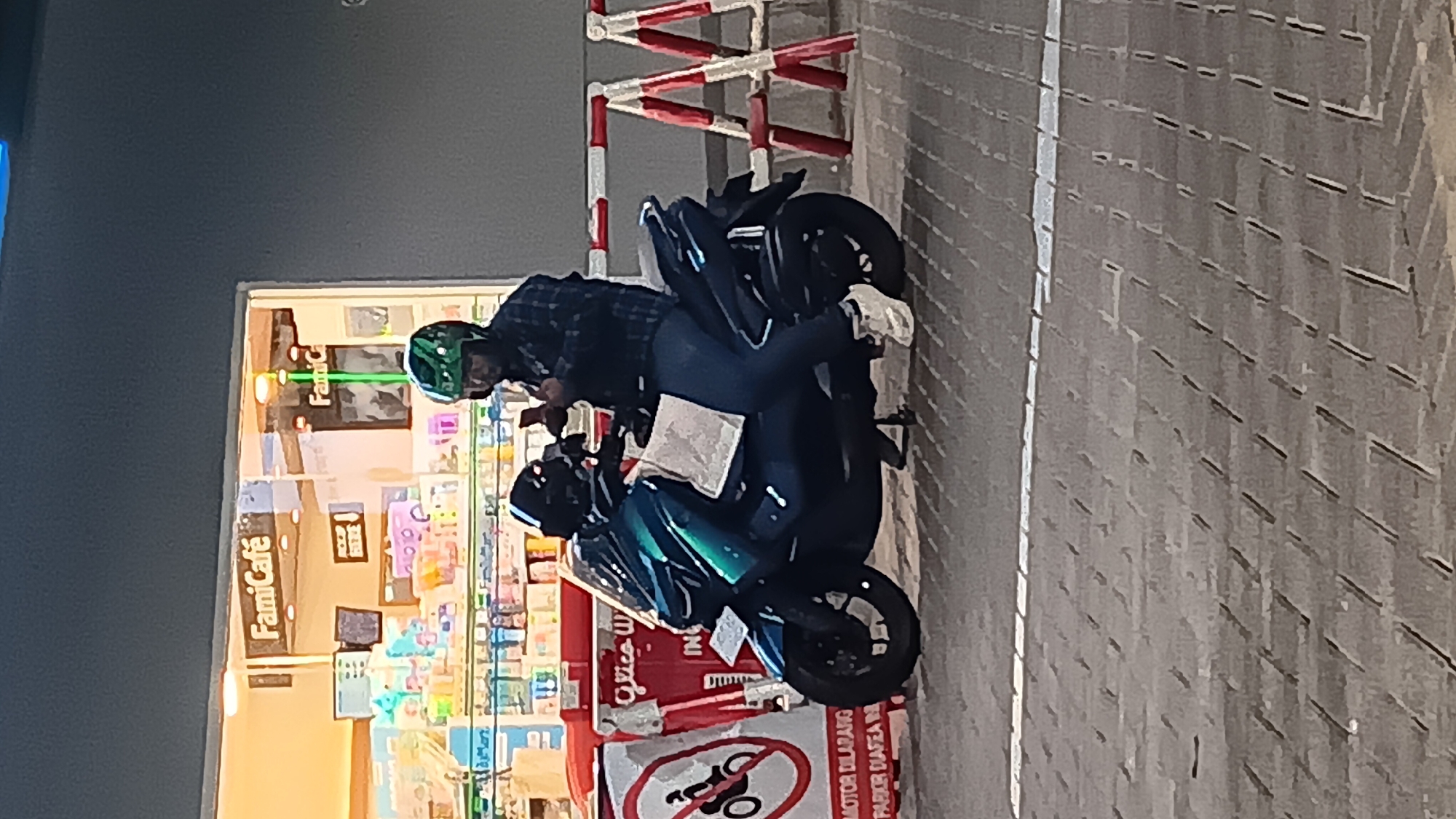 A person wearing a green helmet and a plaid shirt is sitting on a dark blue scooter outside a FamiCafe in South Jakarta, Indonesia. The person is holding a smartphone in their right hand and looking at it. The scooter has a large windshield and a paper bag on the seat. In the background, there are shelves stocked with various items inside the FamiCafe. Red and white barricades are set up in front of the cafe. The ground is paved with interlocking concrete blocks.