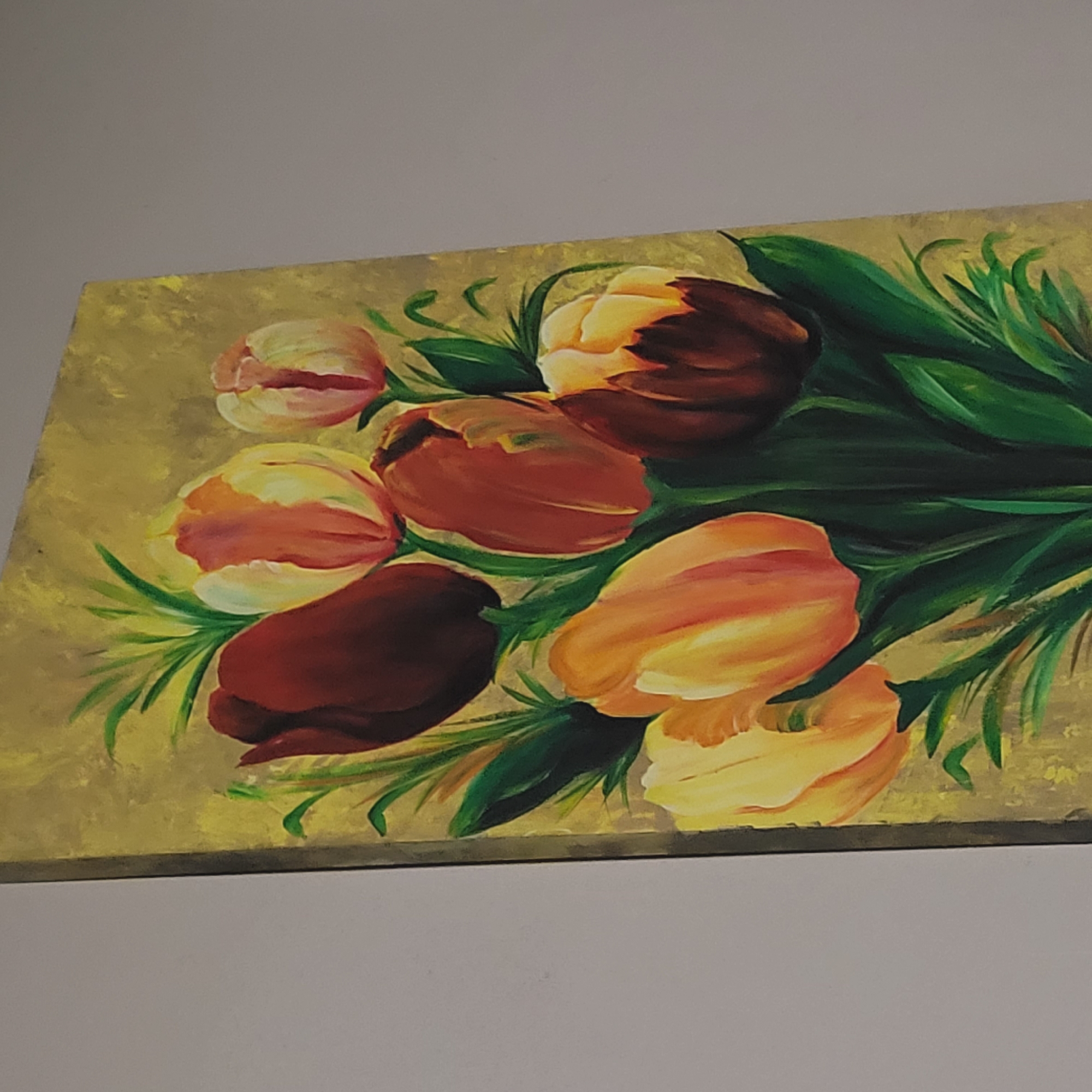 A painting of tulips on a canvas is displayed. The tulips are depicted in shades of red, orange, and yellow with green foliage. The background of the painting is a muted yellow. The painting is located in the city of San Fernando del Valle de Catamarca, Argentina.