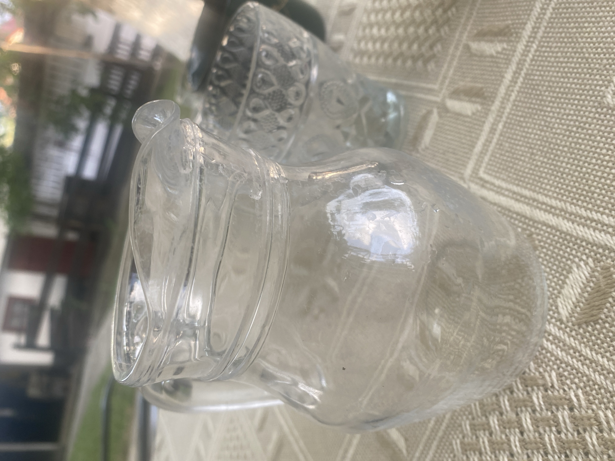 The image depicts a close-up view of a clear glass pitcher with a handle and a spout, placed on a textured, light-colored tablecloth. The pitcher has a rounded body and a reinforced rim. Next to it, partially visible, is a clear glass with an embossed pattern. In the background, out of focus, are elements of an outdoor setting with greenery and a building with red accents, possibly a house. The image does not provide any specific clues to its location within Târgu Neamț, Romania. No people are visible in the image, and no actions or interactions are taking place. The scene appears to be a still life arrangement on a table.