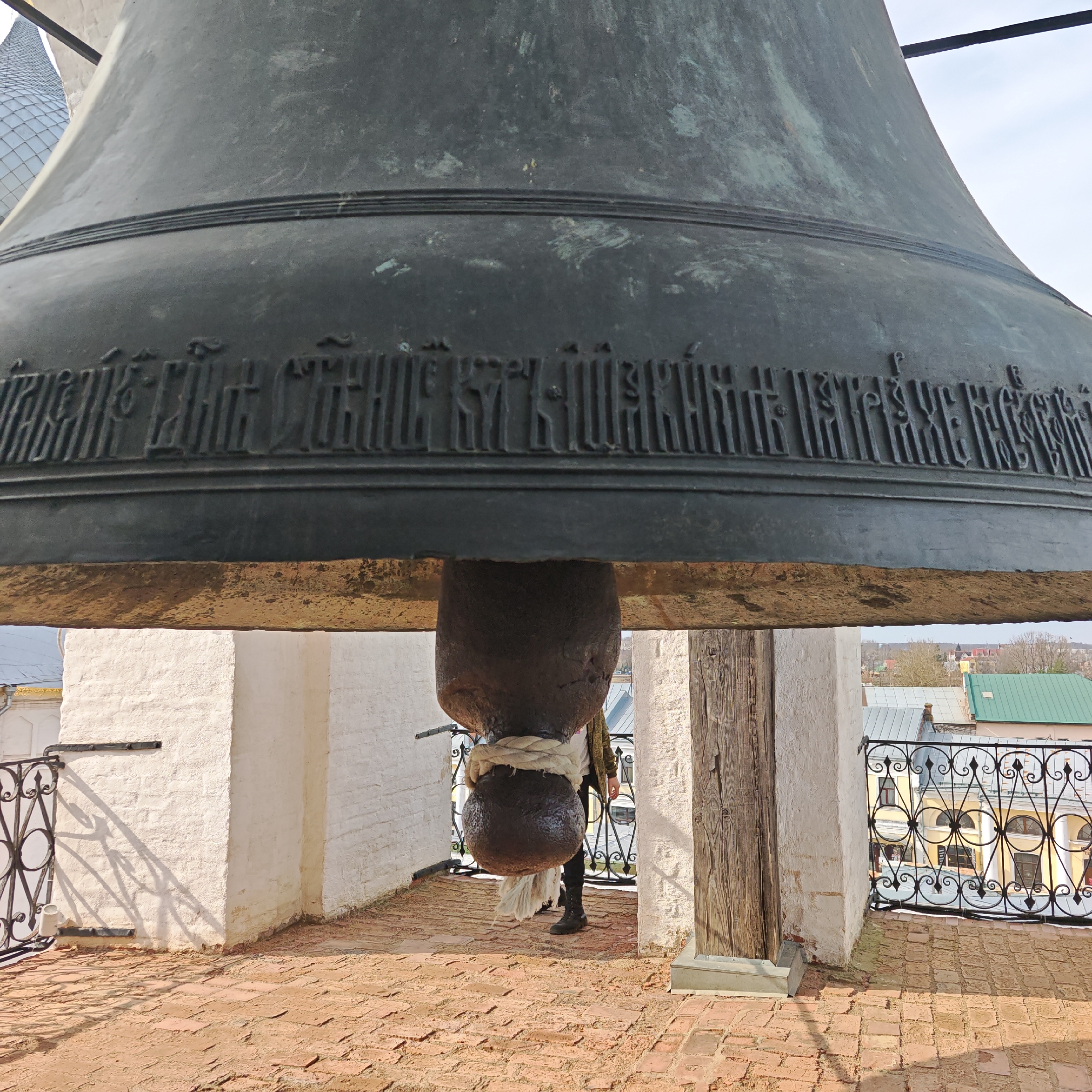 The image shows a large bronze bell suspended in a bell tower, likely in Rostov, Russia. The bell has an inscription around its lower edge in an old Cyrillic script. Below the bell is the clapper, attached with a thick rope, and a person is partially visible. The bell tower features brick flooring, white brick walls, and wooden support beams. The foreground features wrought-iron railings. In the background, there are rooftops and buildings suggesting a town or city. The scene appears to be outdoors, on a bright day.