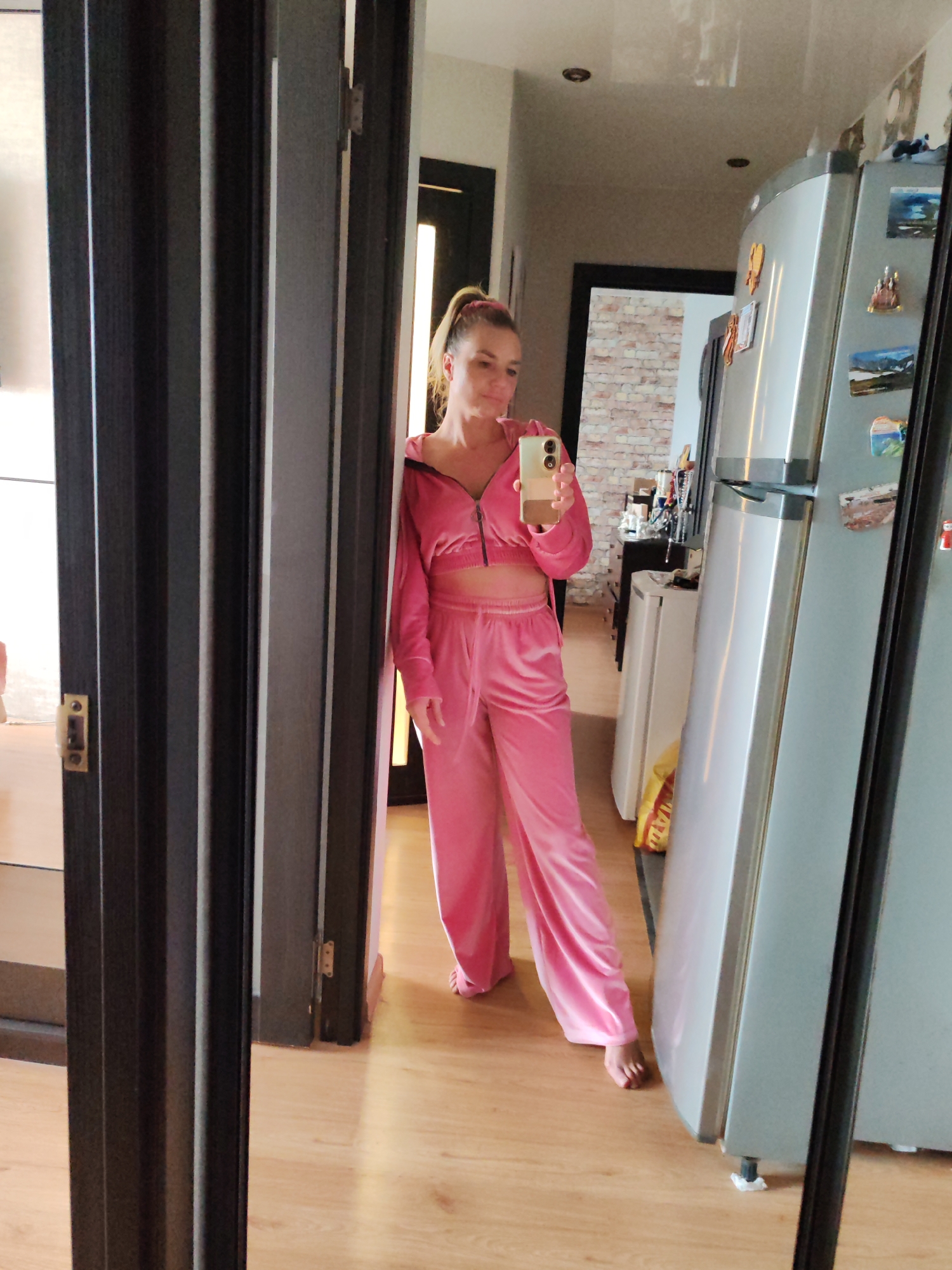 A woman wearing a pink velour tracksuit is standing in an interior space, visible in a mirror's reflection. The woman is fair-skinned, with blonde hair pulled back into a ponytail. She is holding a smartphone in her right hand, seemingly taking a photo of herself in the mirror. The tracksuit consists of a cropped zip-up jacket and wide-leg pants. The room features wooden laminate flooring and is relatively well-lit. A refrigerator with magnets on it is visible to the right, while an open doorway leads to a room with exposed brick and various household objects, including a smaller refrigerator. A folding door is on the left. Given the common interior features, it is not possible to conclusively determine if the location is in Petropavlovsk-Kamchatsky, Russia. There is nothing to indicate this specific location.