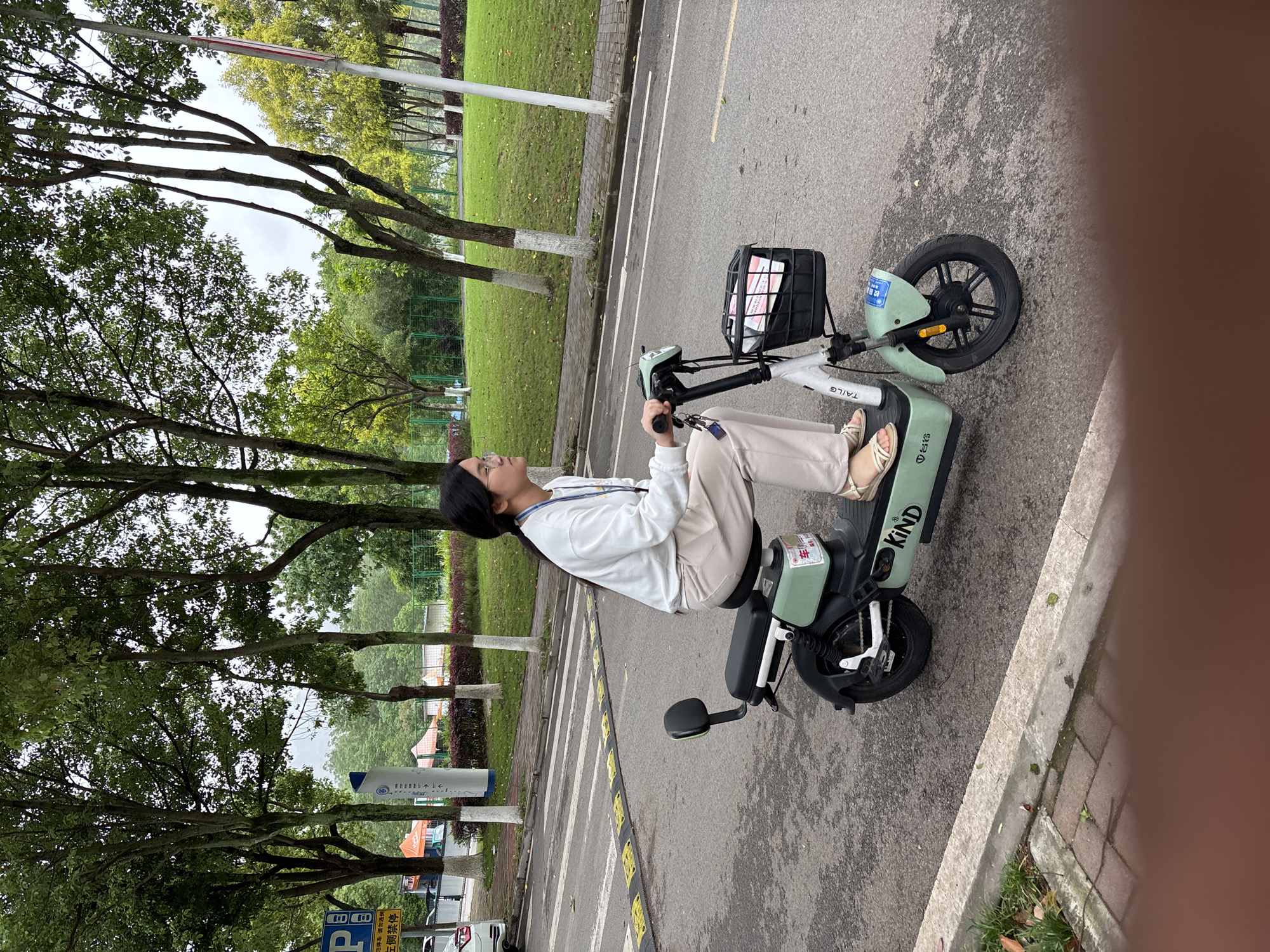A person is sitting on a light green electric scooter on the street in Jiangjin, China. The scooter has a basket in the front. The street is lined with trees and a grassy area. A parking sign is also visible near the trees.
