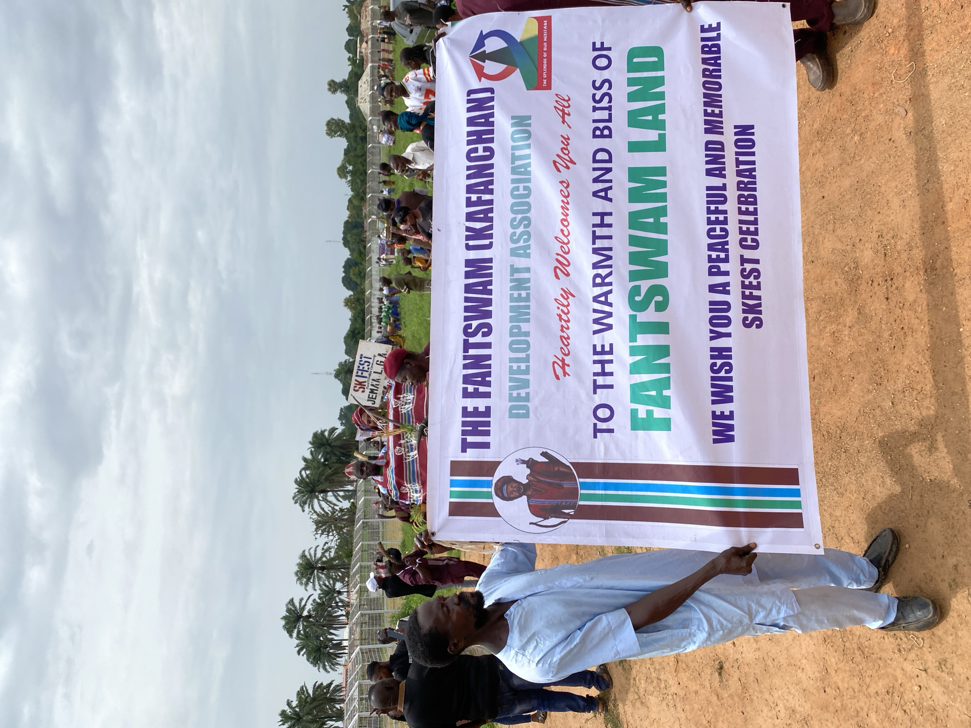 A man in a light blue shirt and dark pants holds up a banner that reads "The Fantswam (Kafanchan) Development Association Heartily Welcomes You All To The Warmth and Bliss of Fantswam Land. We Wish You A Peaceful and Memorable SKFEST Celebration." The banner is being held up in a field surrounded by trees and people. This is in Kafanchan, Nigeria.