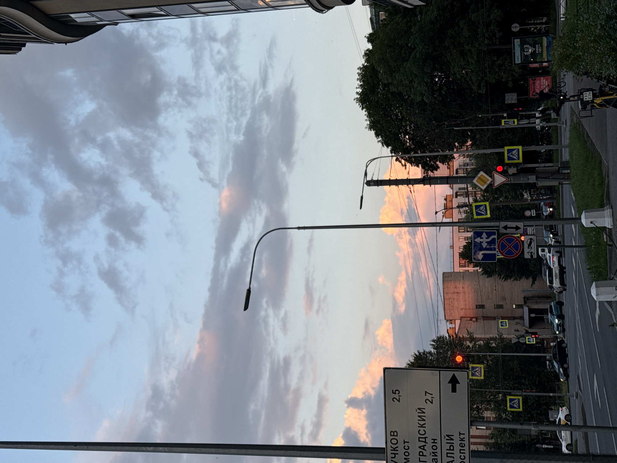 The image depicts an urban scene in Saint Petersburg, Russia, during twilight. The sky is a mix of pale blue and soft pink, with scattered clouds. A tall, dark pole is visible on the left side of the frame, likely a lamppost or utility pole. Further into the scene, a curved streetlamp with a single light fixture arches over the roadway.
Road signs are prominent. One sign, with Cyrillic text and directional arrows, indicates distances to locations such as "Вучков мост" (Vuchkov Bridge) at 2.5 kilometers and "Градский район" (Gradskiy District) at 2.7 kilometers. Another sign points to "Альй проспект" (Aliy Prospect). Multiple pedestrian crossing signs are posted, some illuminated by yellow flashing lights. Traffic lights show a red signal.
A multi-lane road occupies the lower portion of the image, with several cars visible. A white taxi and a dark-colored sedan are in the foreground, with other vehicles further down the road. A turquoise SUV is also present. On the right side of the image, a modern building with curved architecture and multiple windows rises into the sky. Dense green trees partially obscure the lower floors of some buildings, and a smaller building with brick detailing is visible in the middle ground. A bicycle is parked on the sidewalk on the right. The overall atmosphere is that of a city winding down as dusk settles.