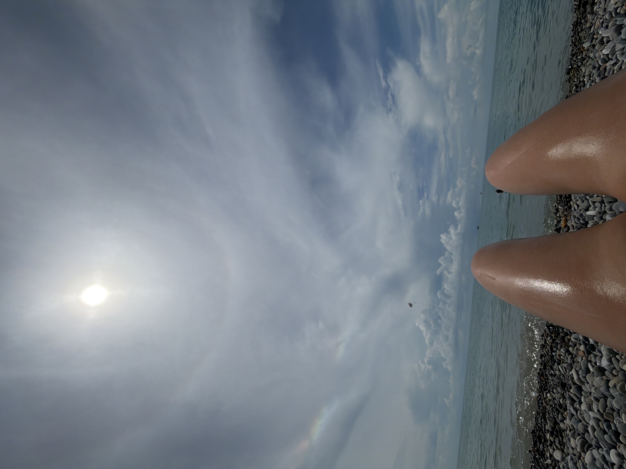 The image depicts a first-person view of a person lying on a pebble beach in Sochi, Russia. The sun is visible high in the sky, creating a halo effect in the clouds. In the distance, a parasailer can be seen airborne. The sea is calm with gentle waves lapping at the shore. The beach is covered with smooth, grey pebbles. The person's legs, oiled and shiny, are prominently featured in the foreground, suggesting a relaxed, sunbathing experience. The overall atmosphere is one of a bright, sunny day at the seaside.