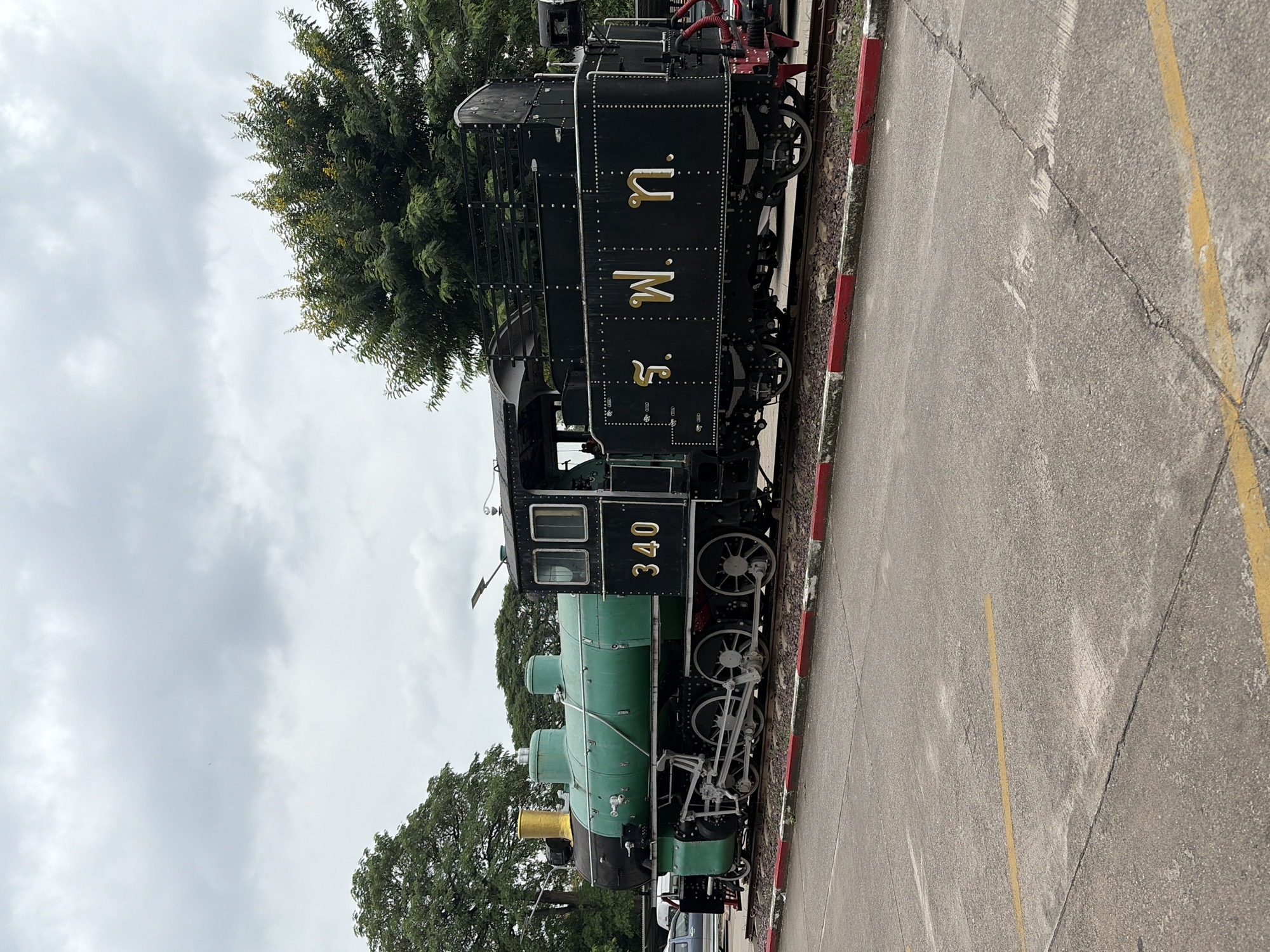 A vintage black and green steam locomotive, numbered 340, is parked on train tracks in Chiang Mai City Municipality, Thailand. The locomotive has a prominent gold chimney. The side of the engine features Thai lettering in gold. The train is situated outdoors on a concrete surface with yellow lines, adjacent to a red and white striped border along the tracks. Trees are visible in the background, and the sky is overcast. There are no people visible in the image.