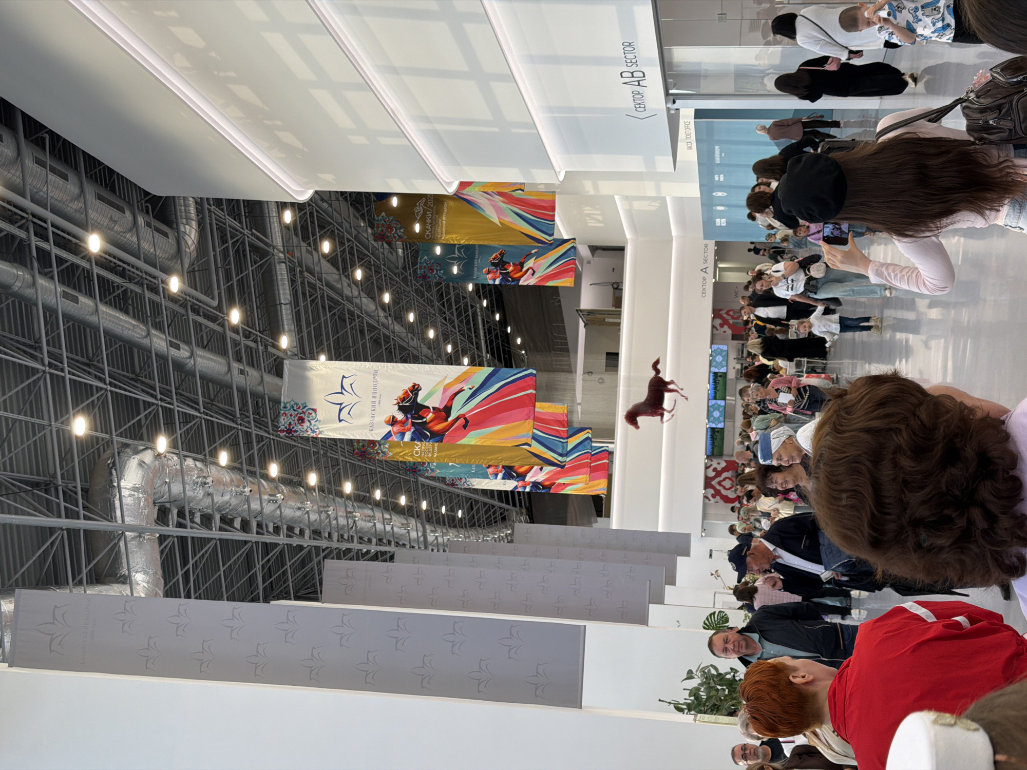 The image depicts a busy indoor space, likely an exhibition hall or arena, with a high ceiling featuring exposed metal structures and ductwork. Several large banners hang from the ceiling, some displaying graphics of horses and jockeys, and others with ornamental patterns. In the foreground, a crowd of people are milling about. Some are looking at displays, while others are engaged in conversation. A woman in the foreground is taking a photo with her phone. The environment is well-lit by overhead lighting. Signs indicating "SECTOR AB" and "SECTOR A" are visible. A model of a horse is suspended from the ceiling in the background.