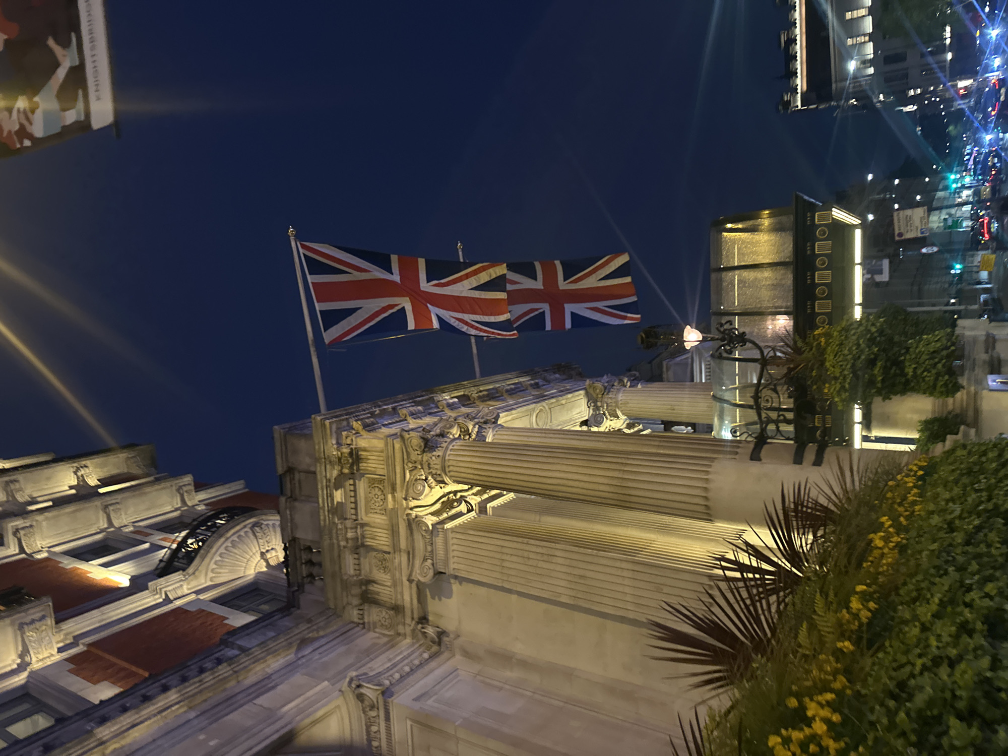The image depicts a nighttime scene in London, United Kingdom. Two British Union Jack flags are prominently displayed atop a classical building with ornate columns. The building appears to be illuminated with spotlights. A banner with the word "KNIGHTSBRIDGE" is partially visible in the upper-left corner of the frame, suggesting the location. A glimpse of the city is seen to the right, with traffic lights and other buildings illuminated. Lush greenery is present in the foreground.